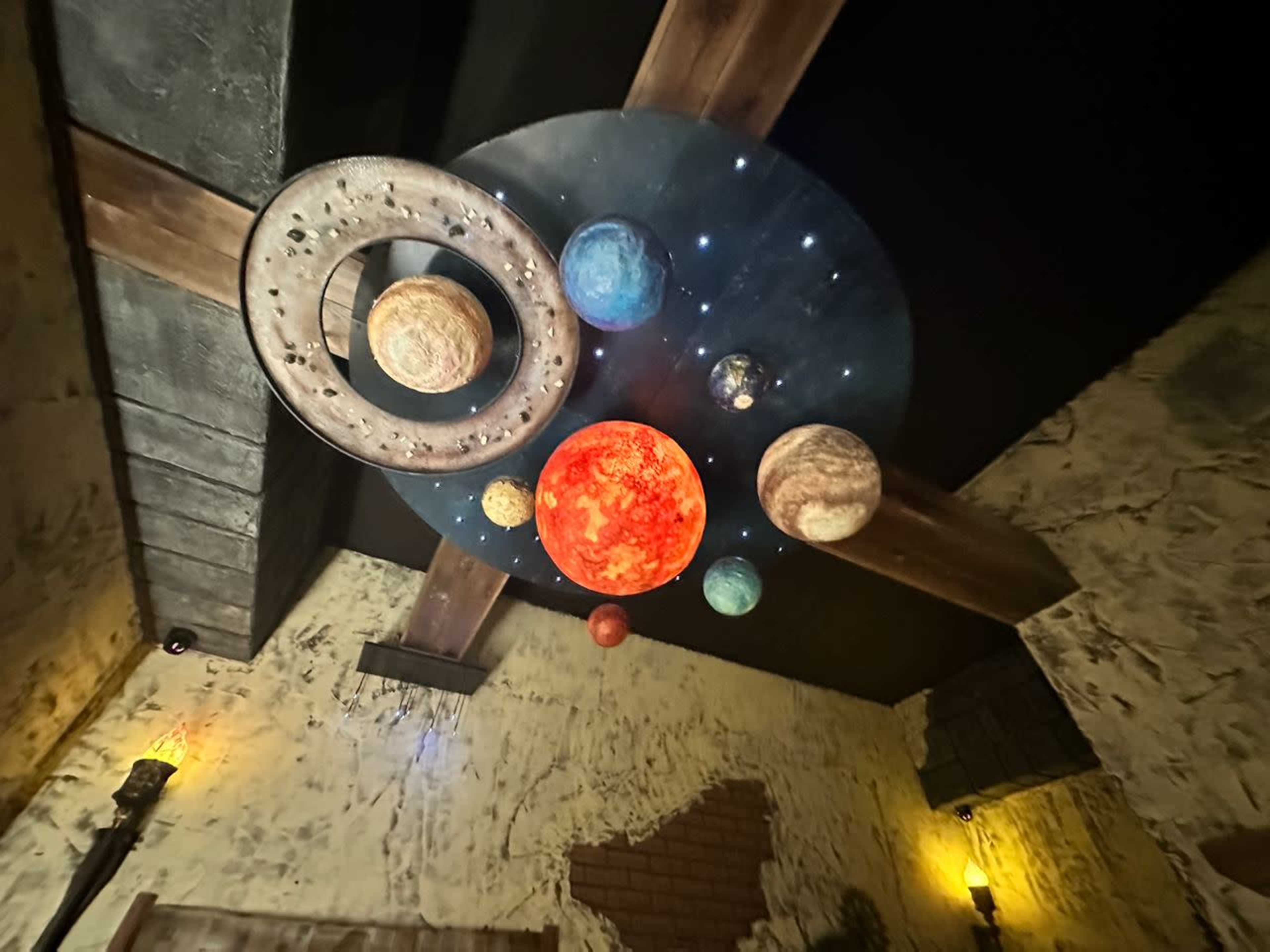 A circular ceiling display features a realistic arrangement of planets and a ringed planet hanging above a room with textured walls and dim lighting.