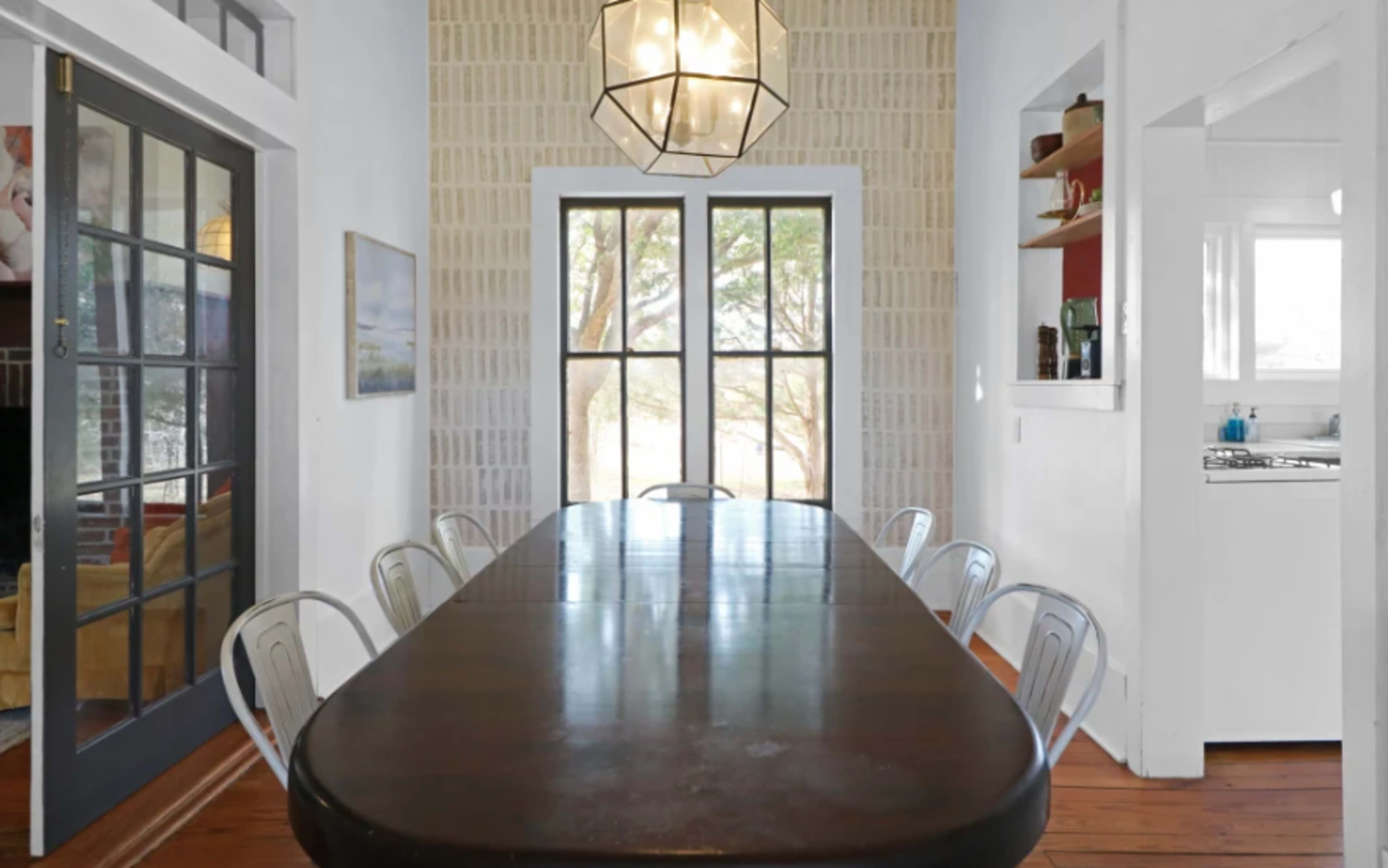 A rectangular wooden dining table is set in a bright room with large windows and a contemporary light fixture overhead.