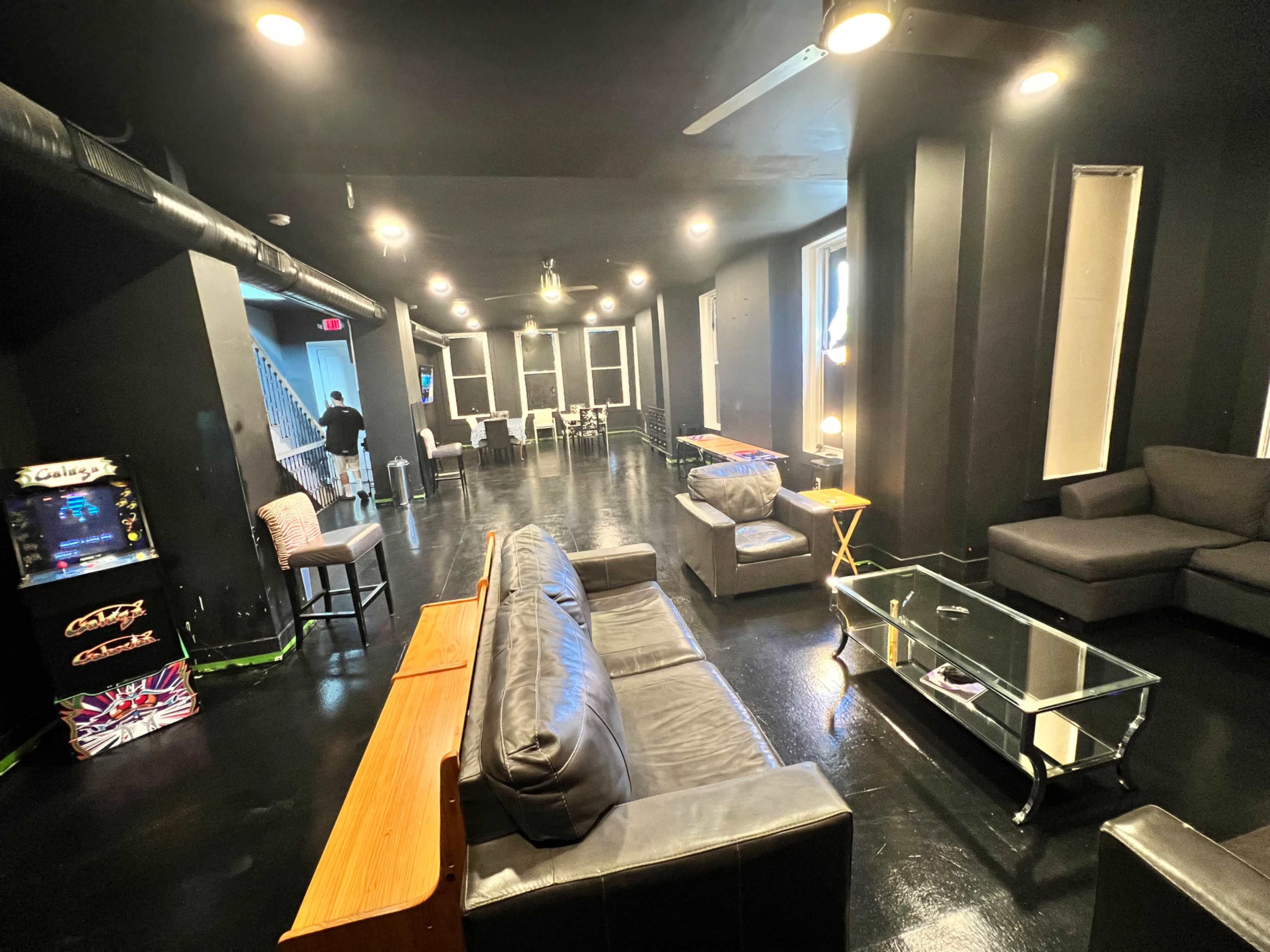 A dimly lit lounge area featuring black walls, several couches, a glass coffee table, and an arcade machine in the corner.