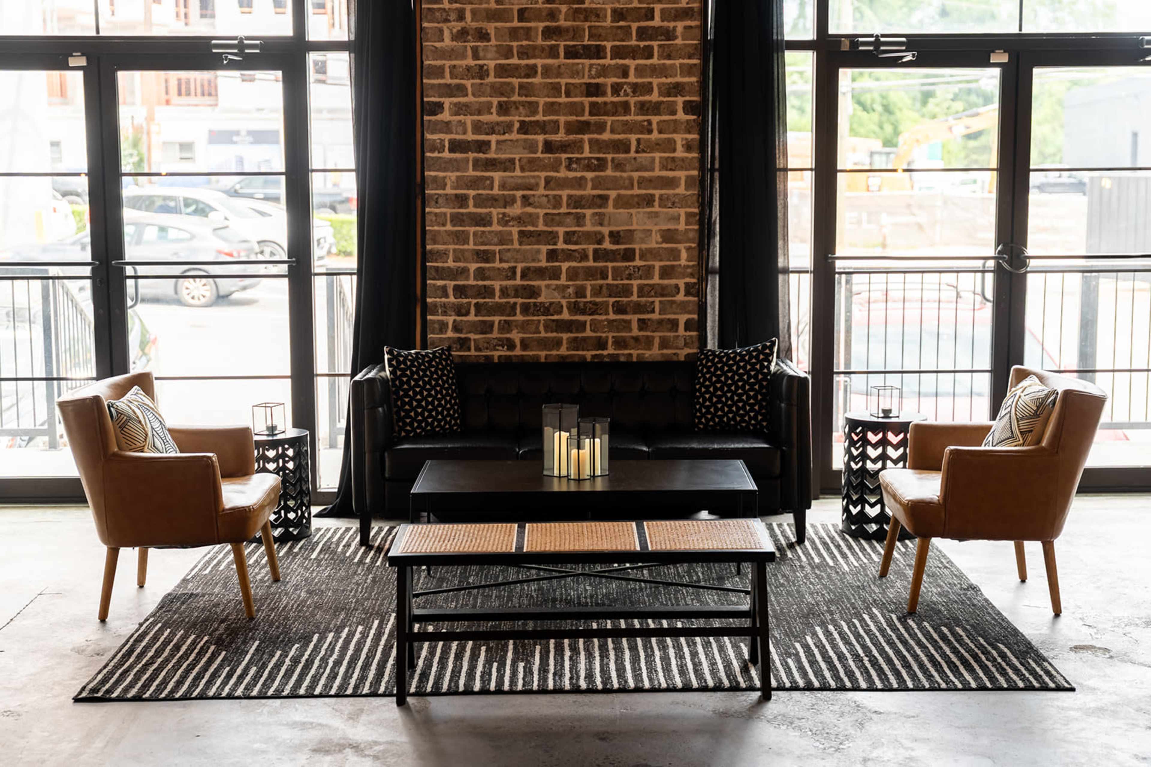 The image shows a stylish living room arrangement featuring a black sofa, two tan armchairs, a coffee table, and decorative elements against a backdrop of large windows and exposed brick walls.