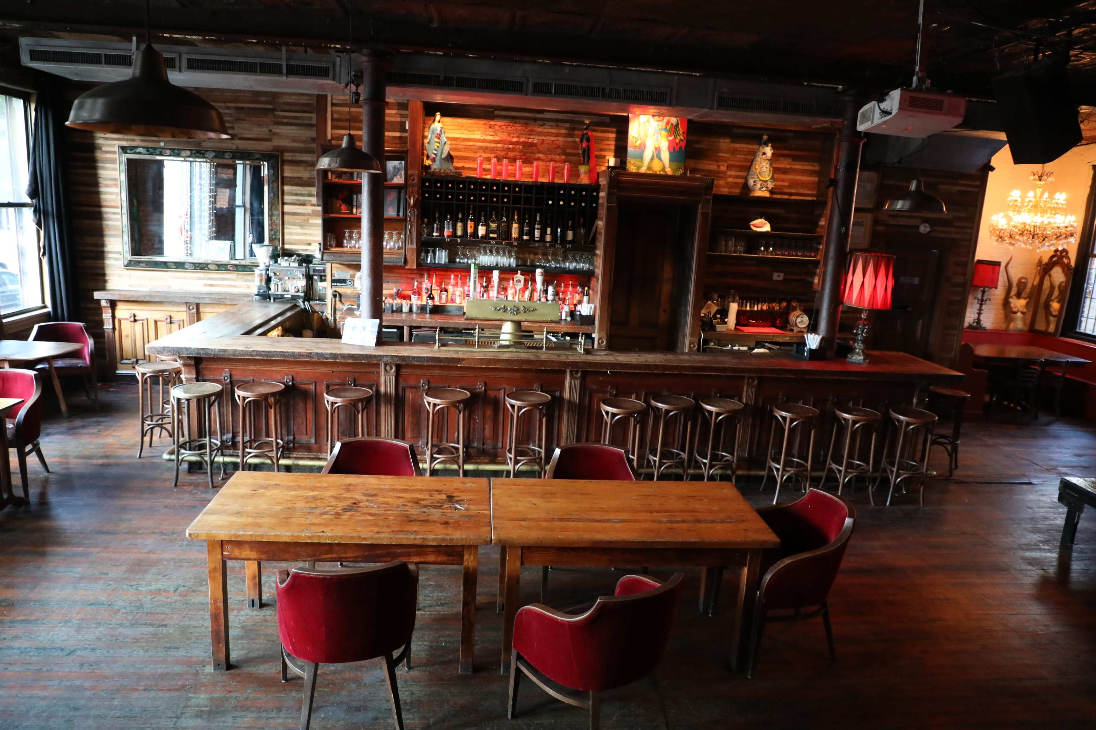 A rustic bar area with a long wooden counter, high stools, and two wooden tables surrounded by red chairs in a dimly lit interior.