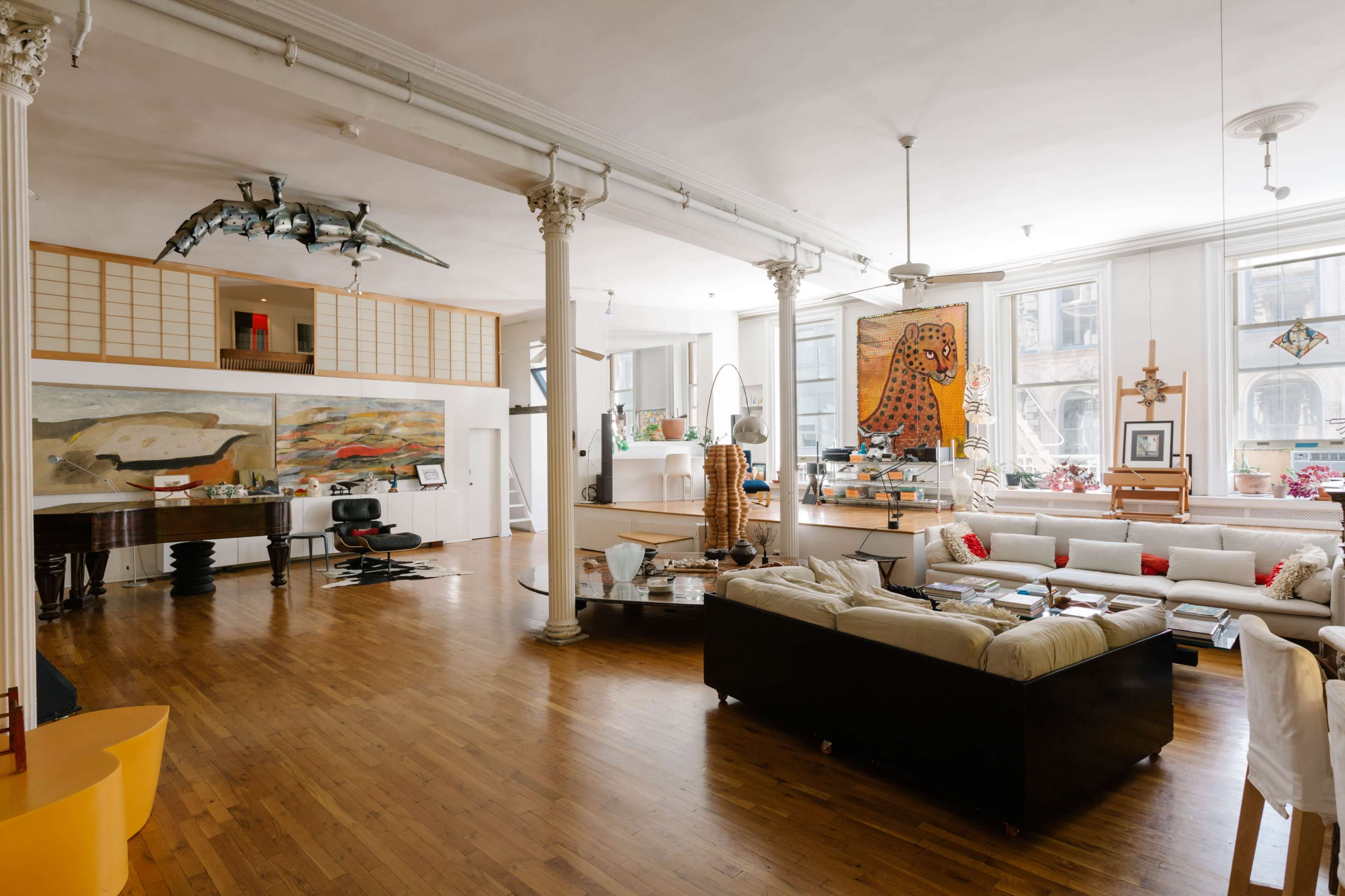 The spacious interior features wooden flooring, tall pillars, and a mix of modern and eclectic furniture, including a grand piano and artwork on the walls.