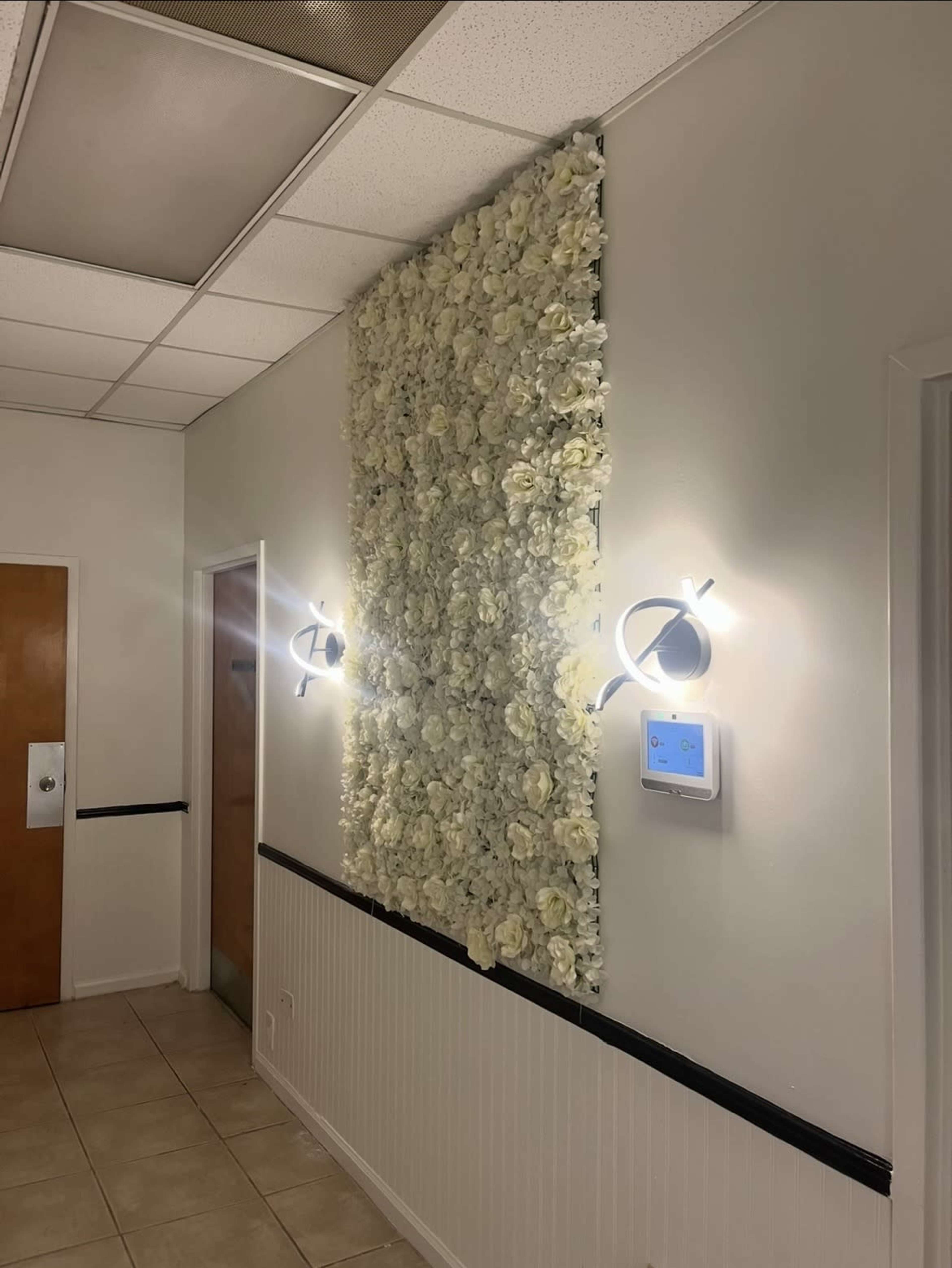 A wall covered with white artificial flowers is illuminated by two wall sconces, beside a control panel in a hallway with light tile flooring.