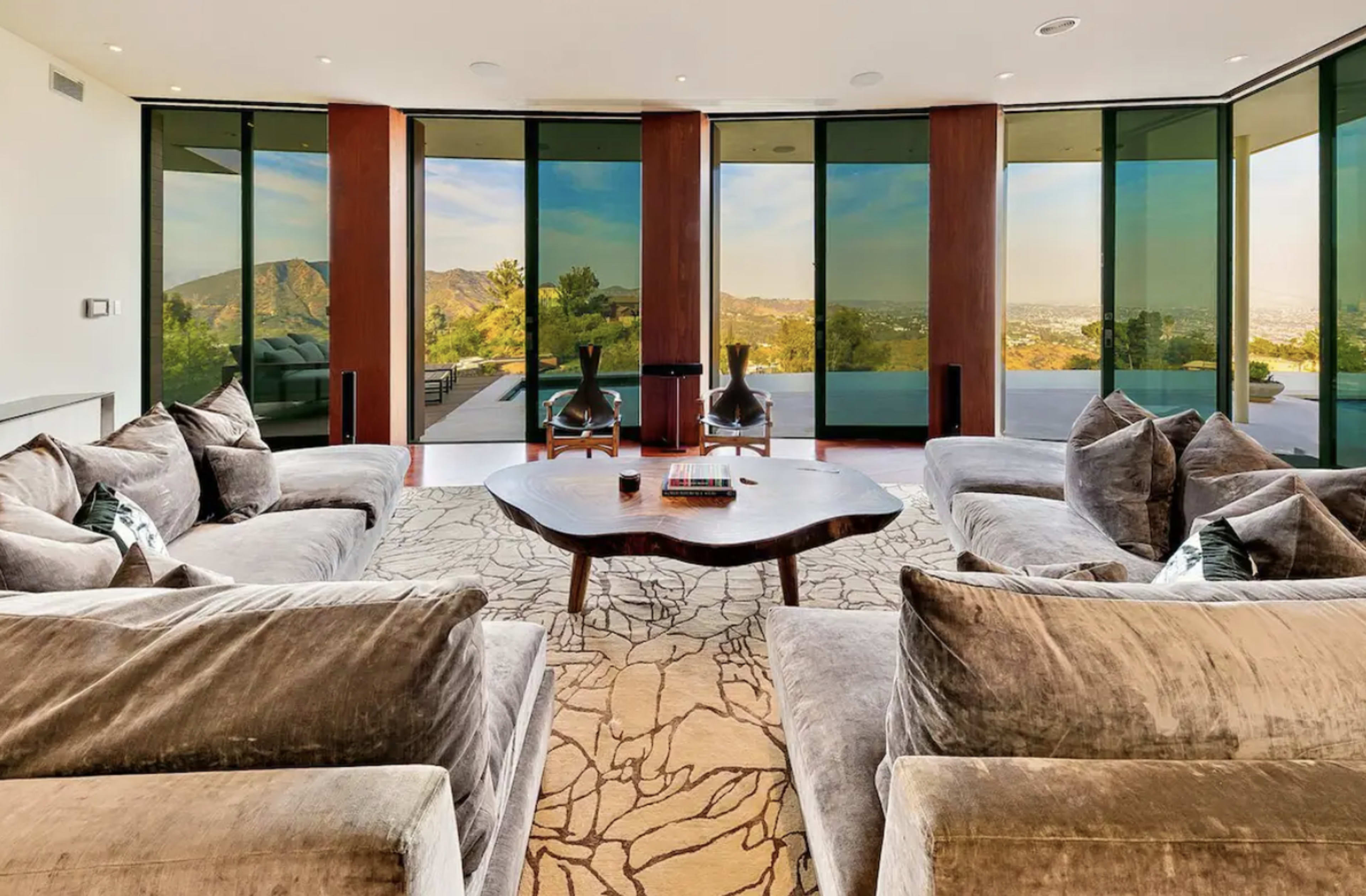 A modern living room with large glass windows overlooking a mountainous view, featuring plush sofas and a wooden coffee table.