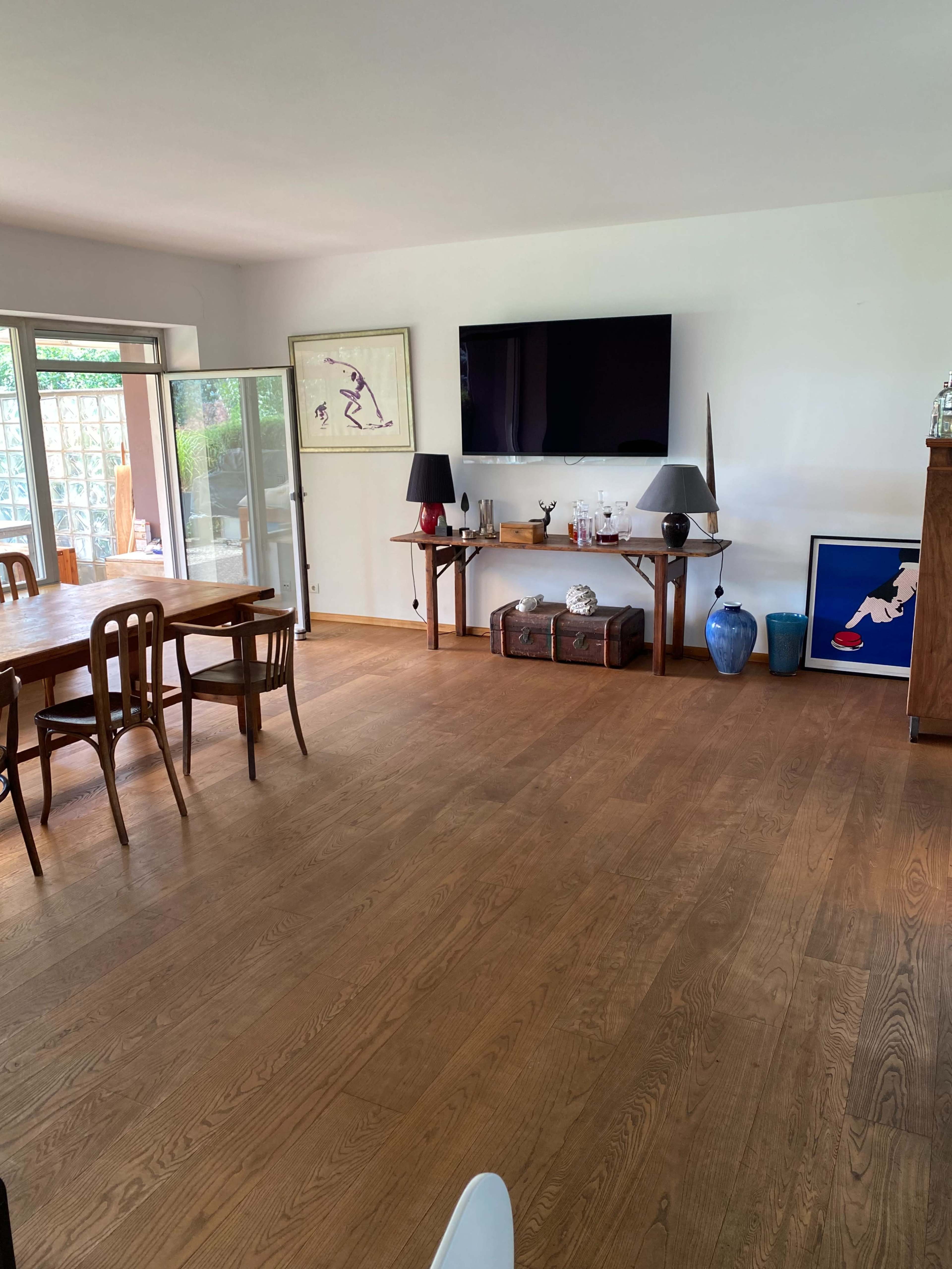 A modern living room with wooden flooring, a large television mounted on the wall, a dining table with chairs, and various decorative items on display.