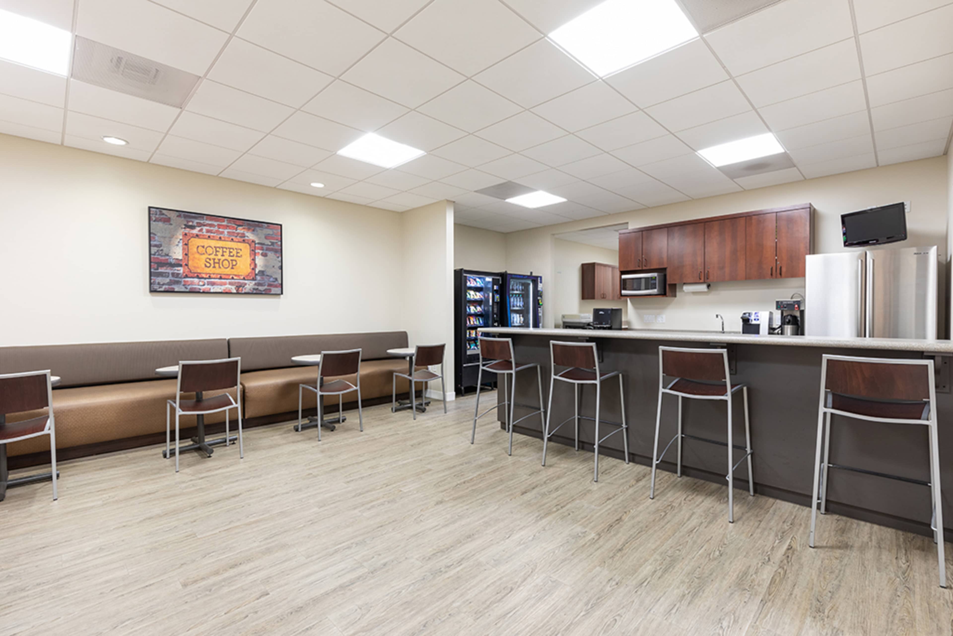 The image shows a modern break room equipped with a coffee shop sign, a kitchenette area, seating, and a vending machine.