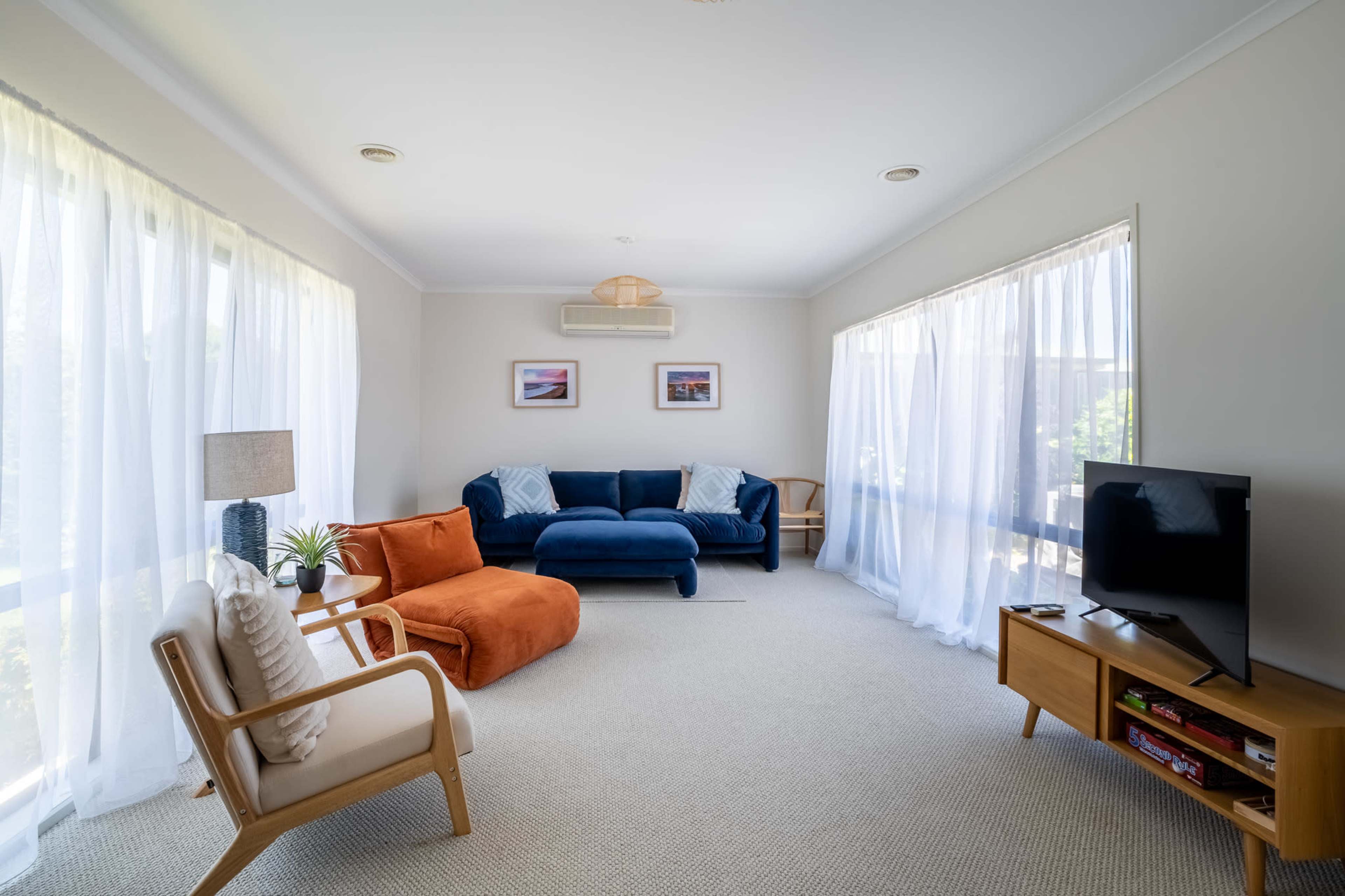 The living room features a blue sofa, an orange lounge chair, a television on a wooden stand, and large windows with sheer white curtains.