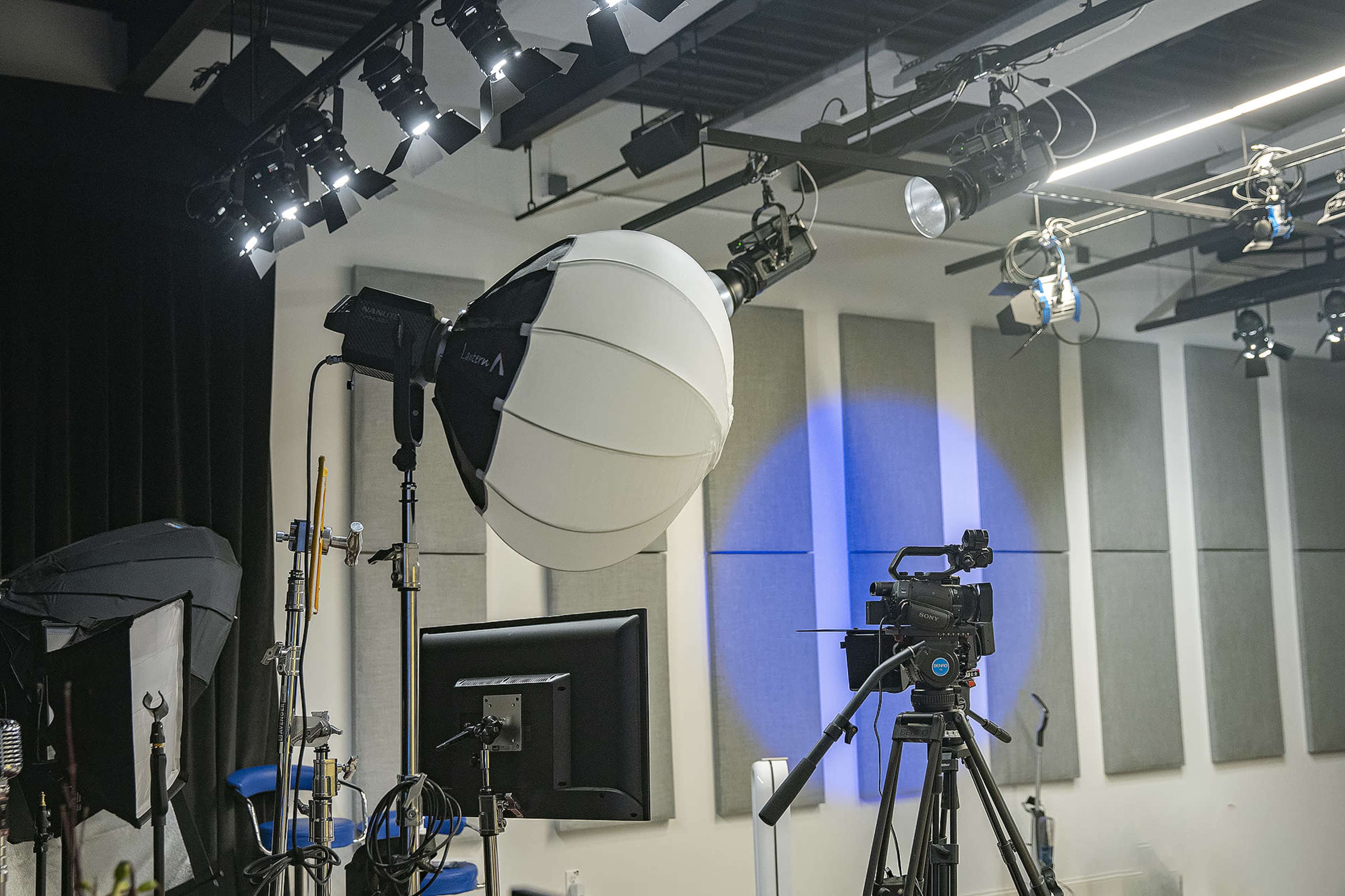 The image shows a photography studio equipped with a large softbox light, a video camera on a tripod, and various lighting equipment mounted on the ceiling.