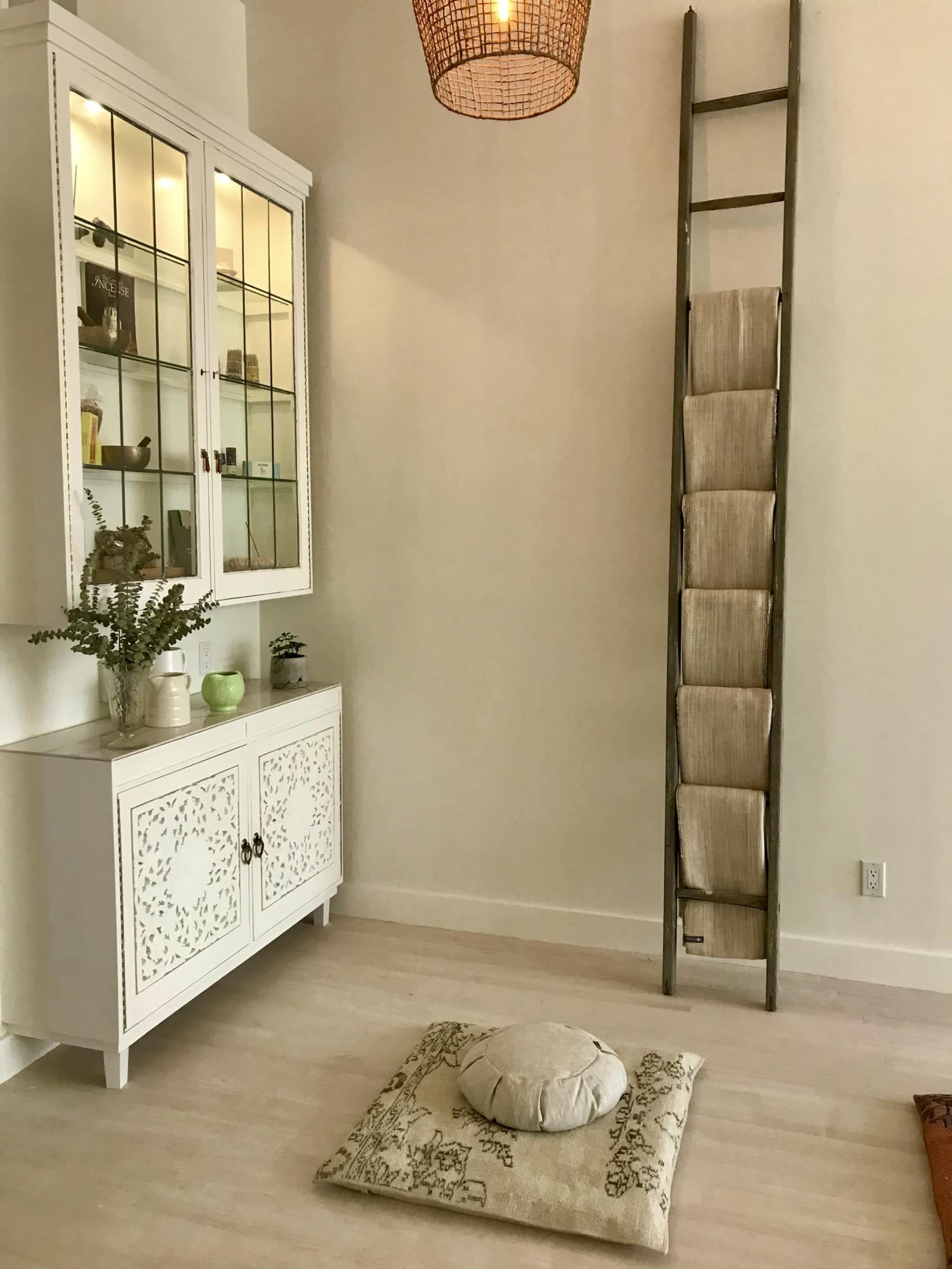 The image shows a serene interior space featuring a white cabinet with glass doors, a decorative ladder holding stacked fabric, and a cushion placed on the floor.