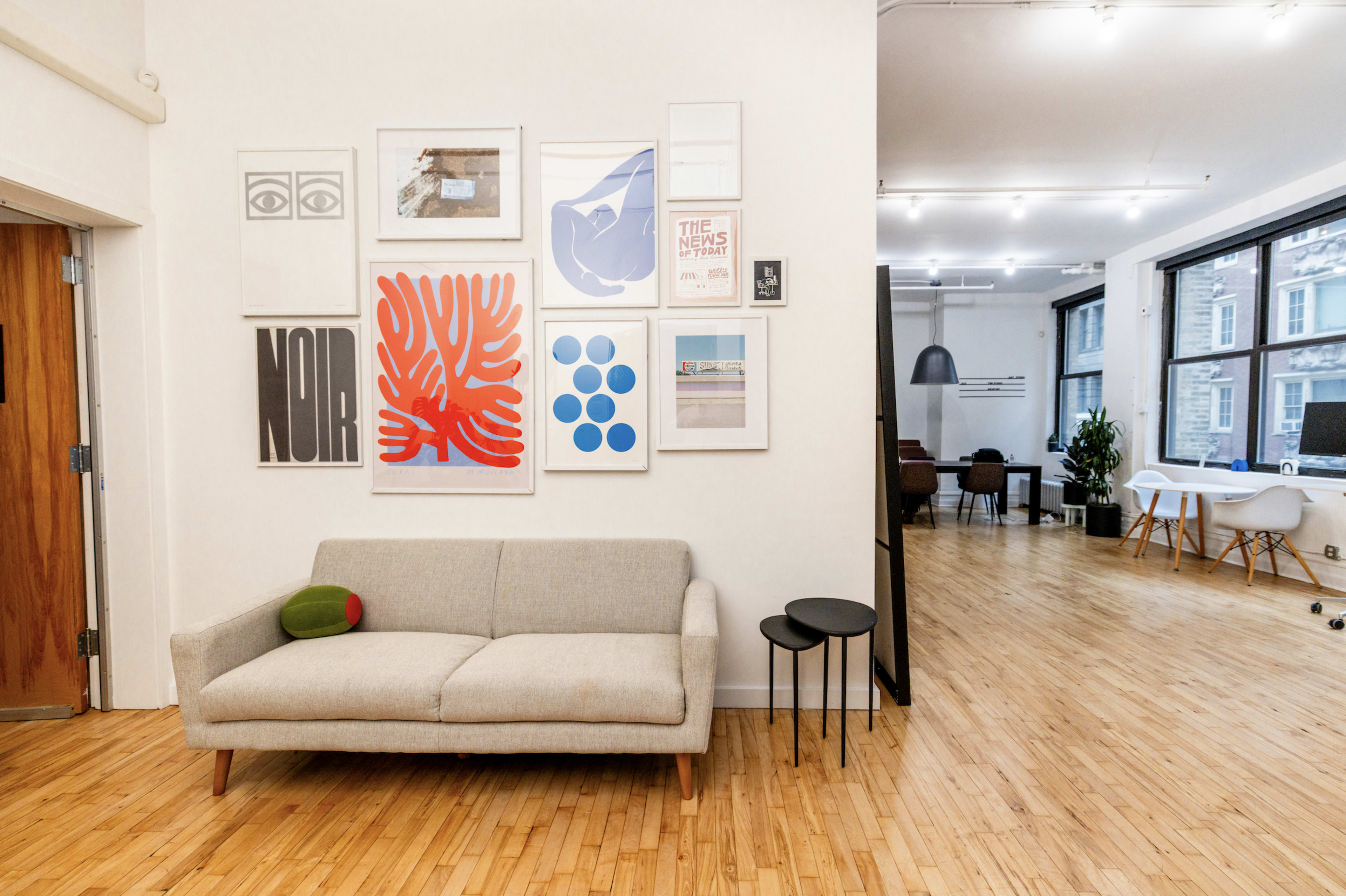 The image shows a modern office interior featuring a gray sofa against a wall adorned with various framed artworks, with a small black side table nearby and an open workspace in the background.
