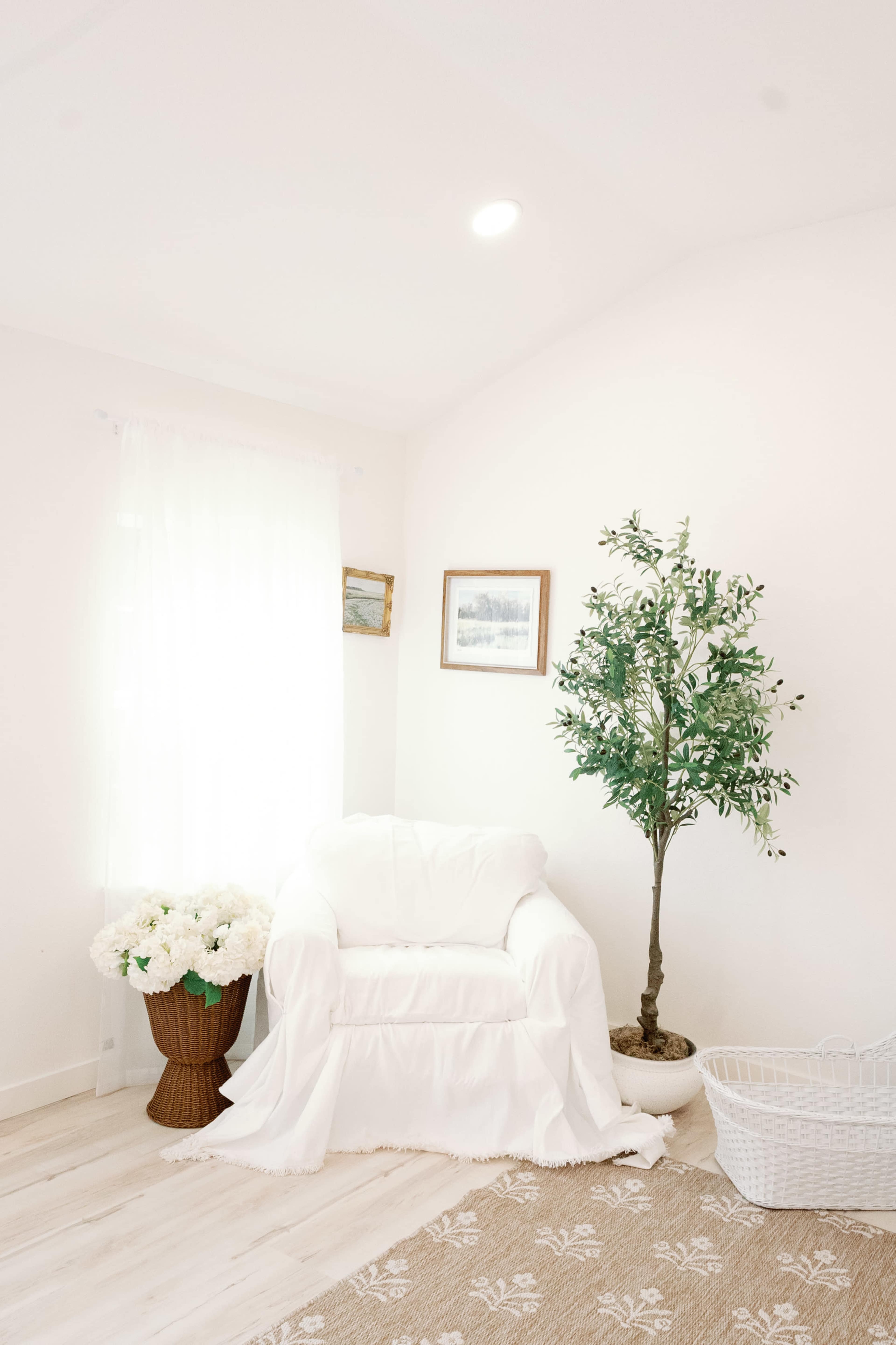 The image shows a bright, minimalist room featuring a white armchair, a potted plant, a basket, and floral arrangements in a wicker container.
