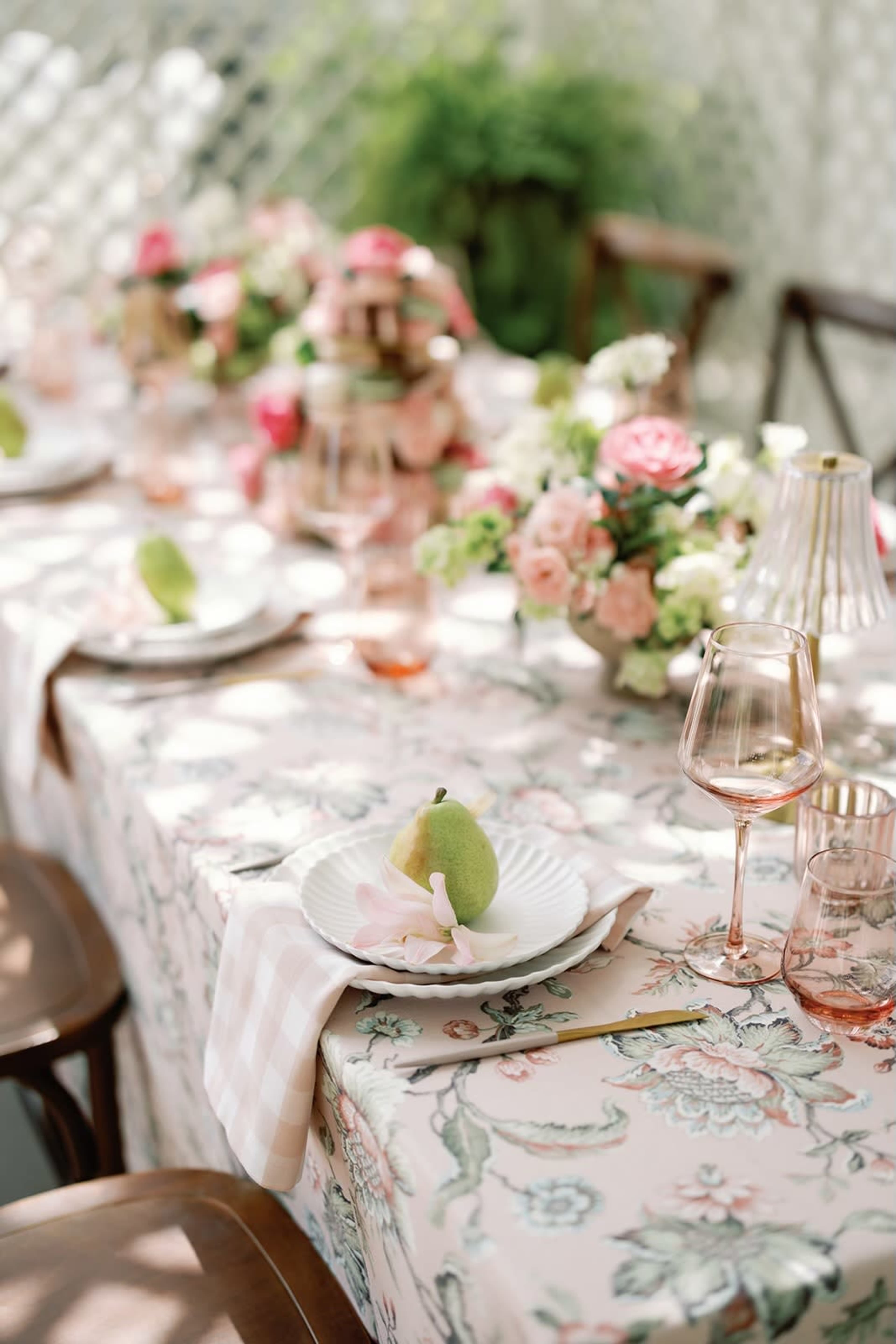 A dining table is elegantly set with floral tablecloths, white plates featuring green pears, and decorative glassware surrounded by floral arrangements.