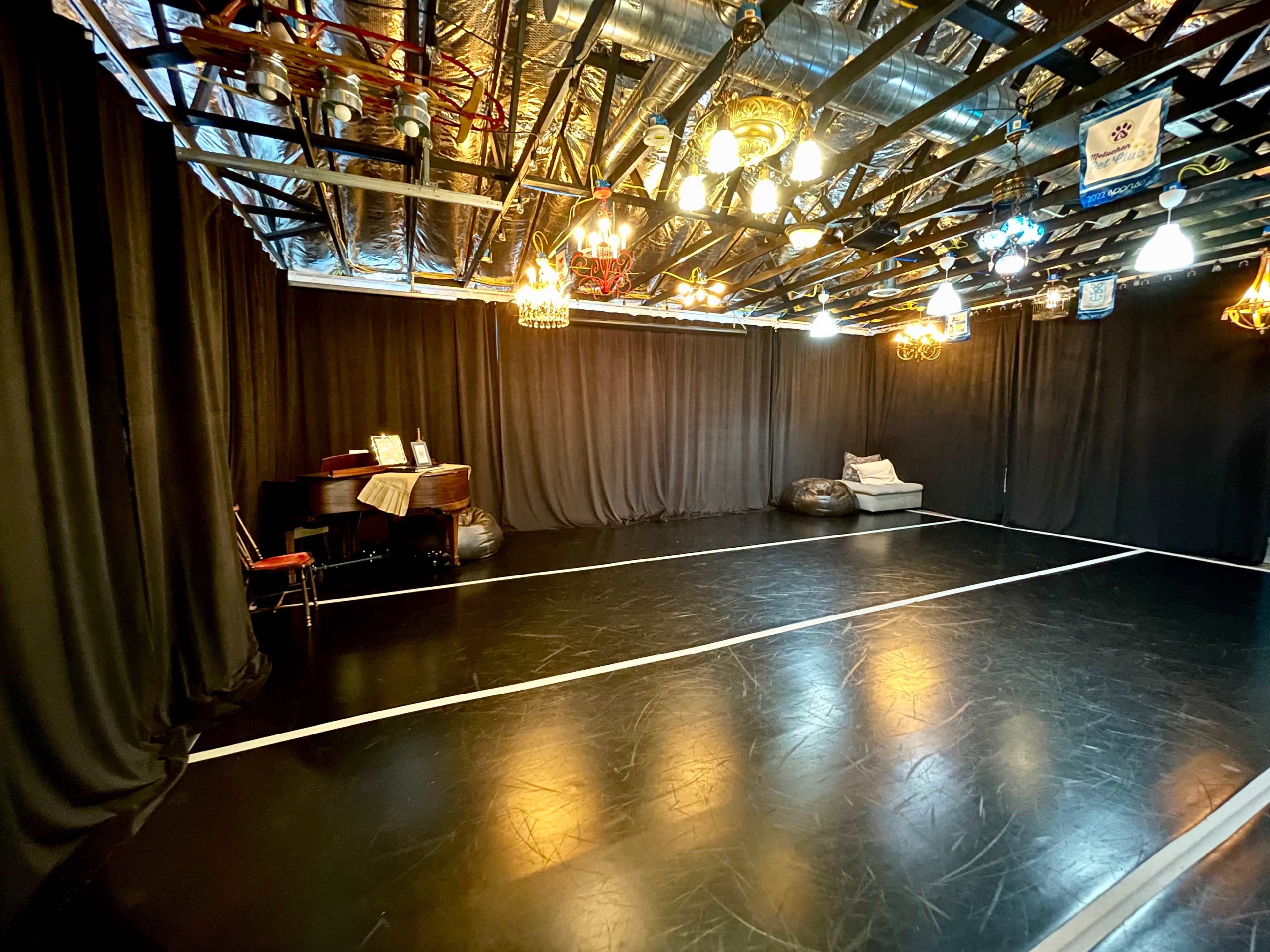 A spacious dance studio features a black floor, mirrored walls, and various hanging light fixtures, with a piano and seating area along one side.