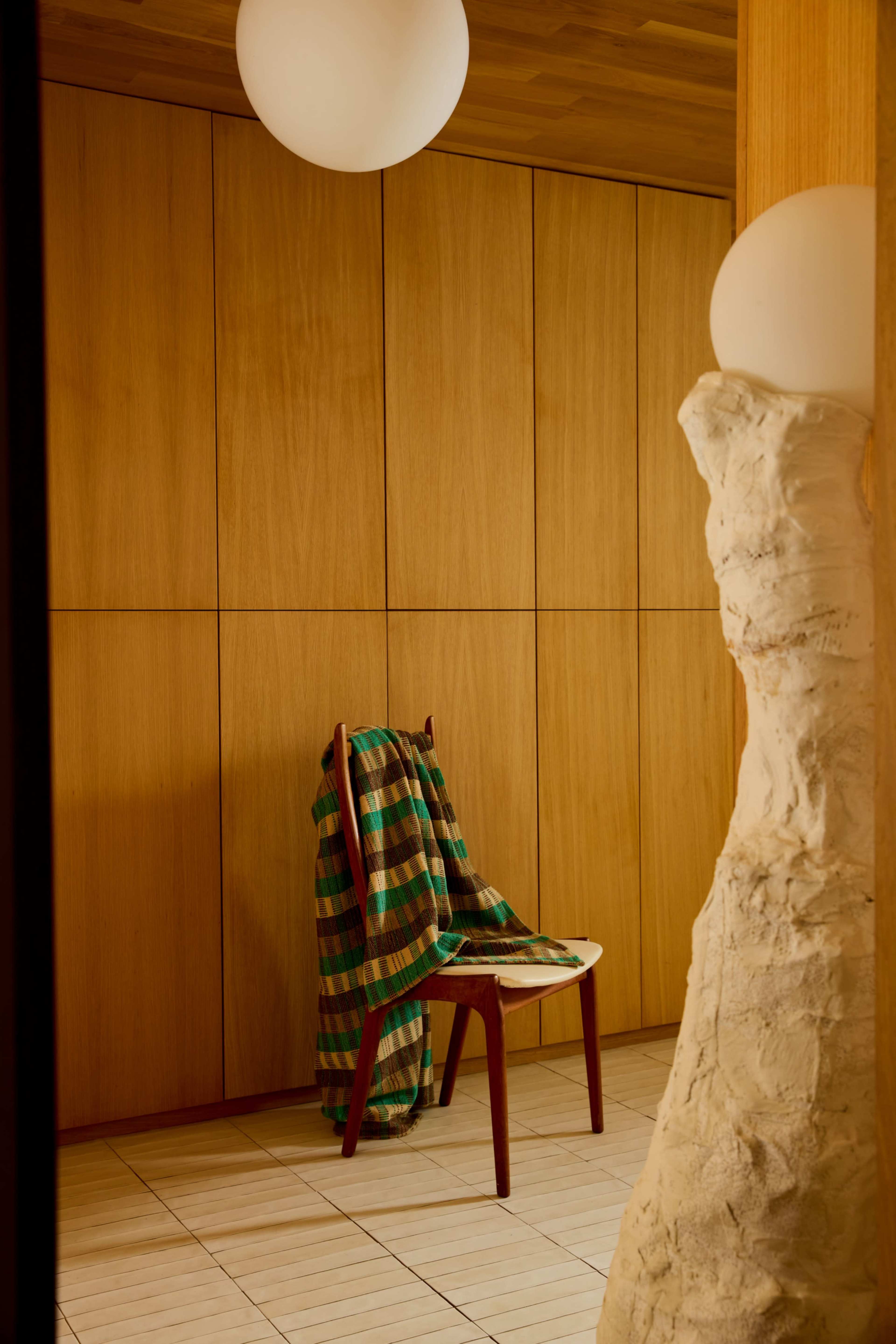 A wooden chair with a colorful blanket draped over it stands against a wall of wooden panels in a minimalist interior space.