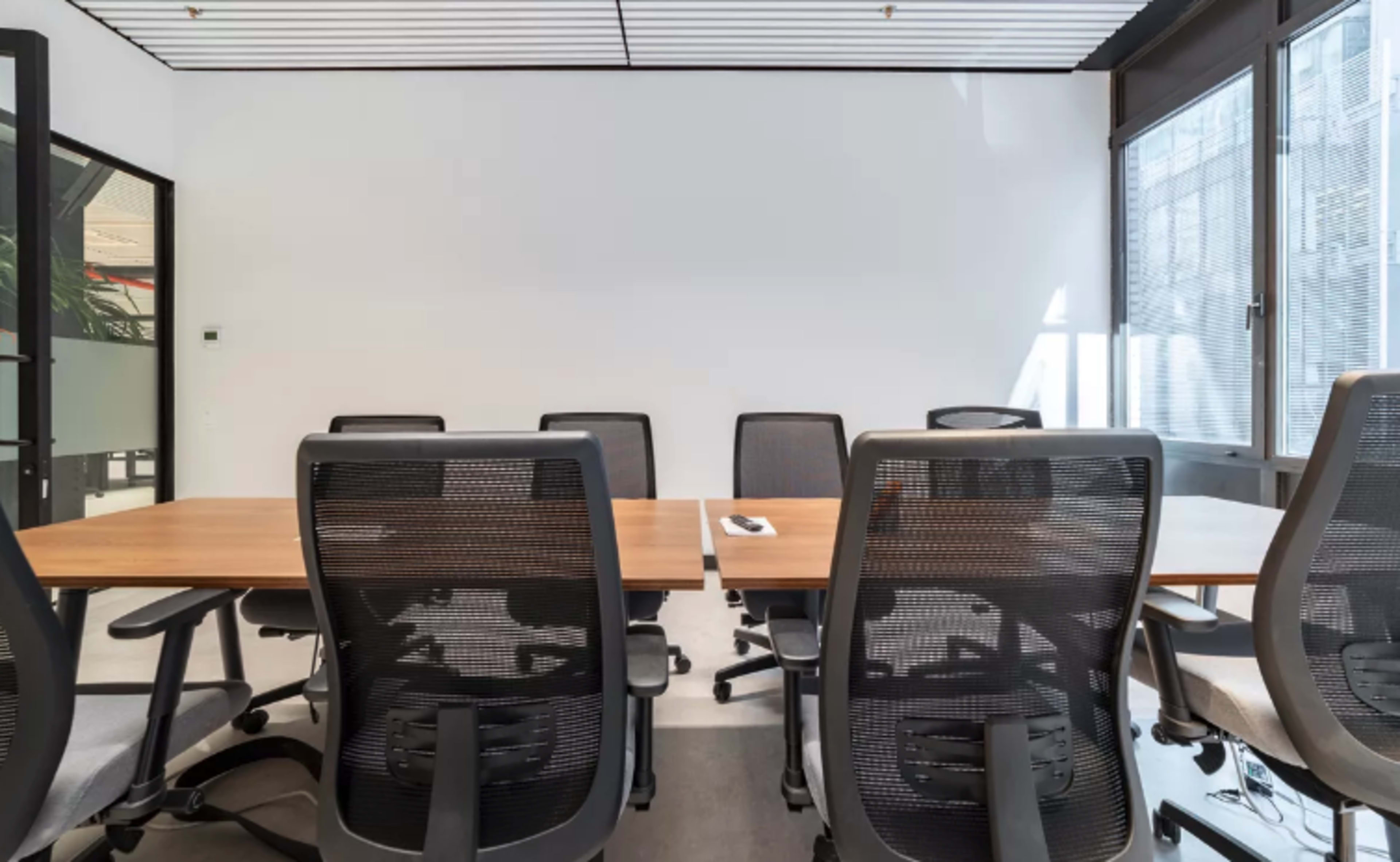 A modern conference room features a wooden table surrounded by ergonomic office chairs, with large windows allowing natural light to fill the space.