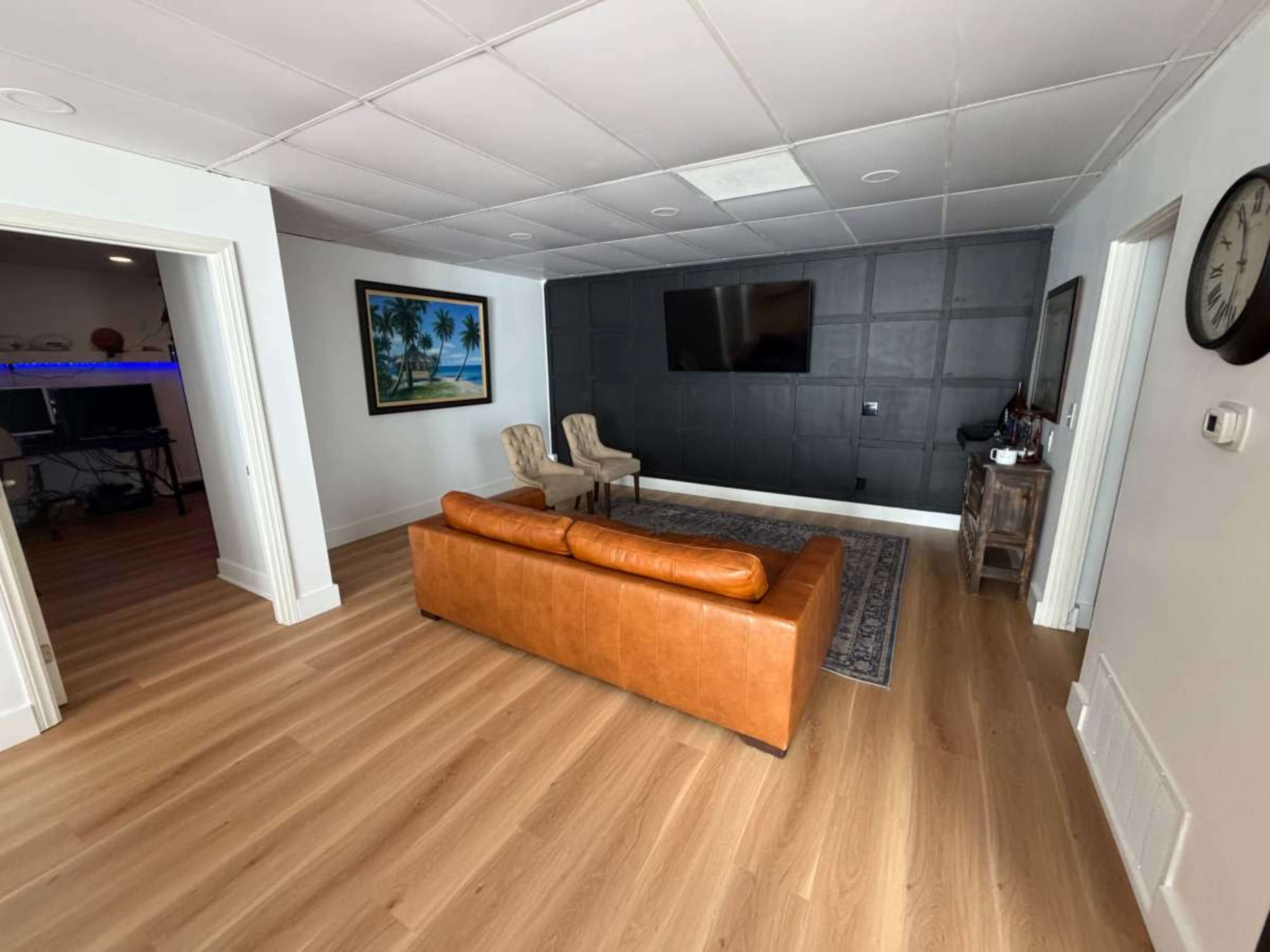 The room features a leather sofa, two armchairs, a wall-mounted television, and hardwood flooring.