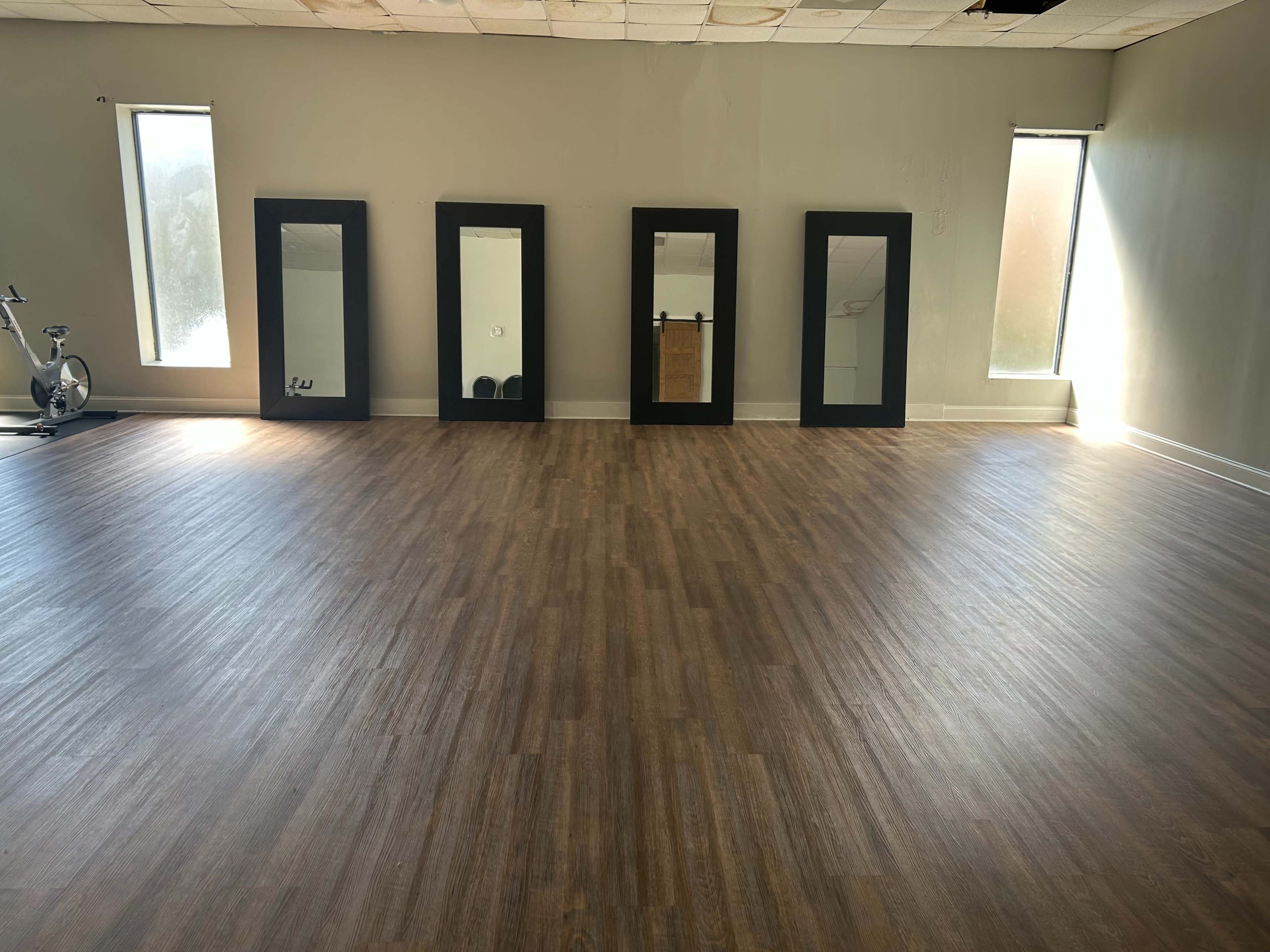 A spacious room with a wooden floor, two windows allowing natural light, and five large mirrors against the wall.