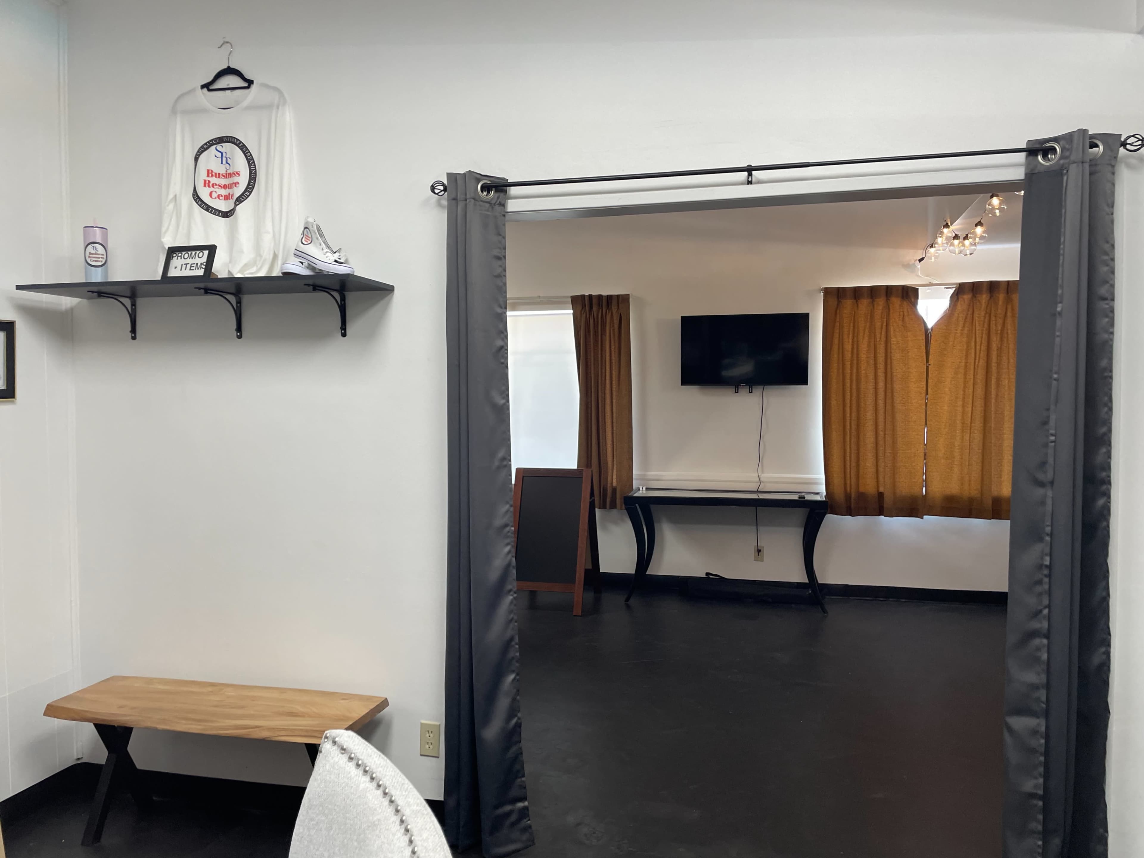 A room with a shelf displaying a shirt and a bench, leading to another room featuring a television and curtains.