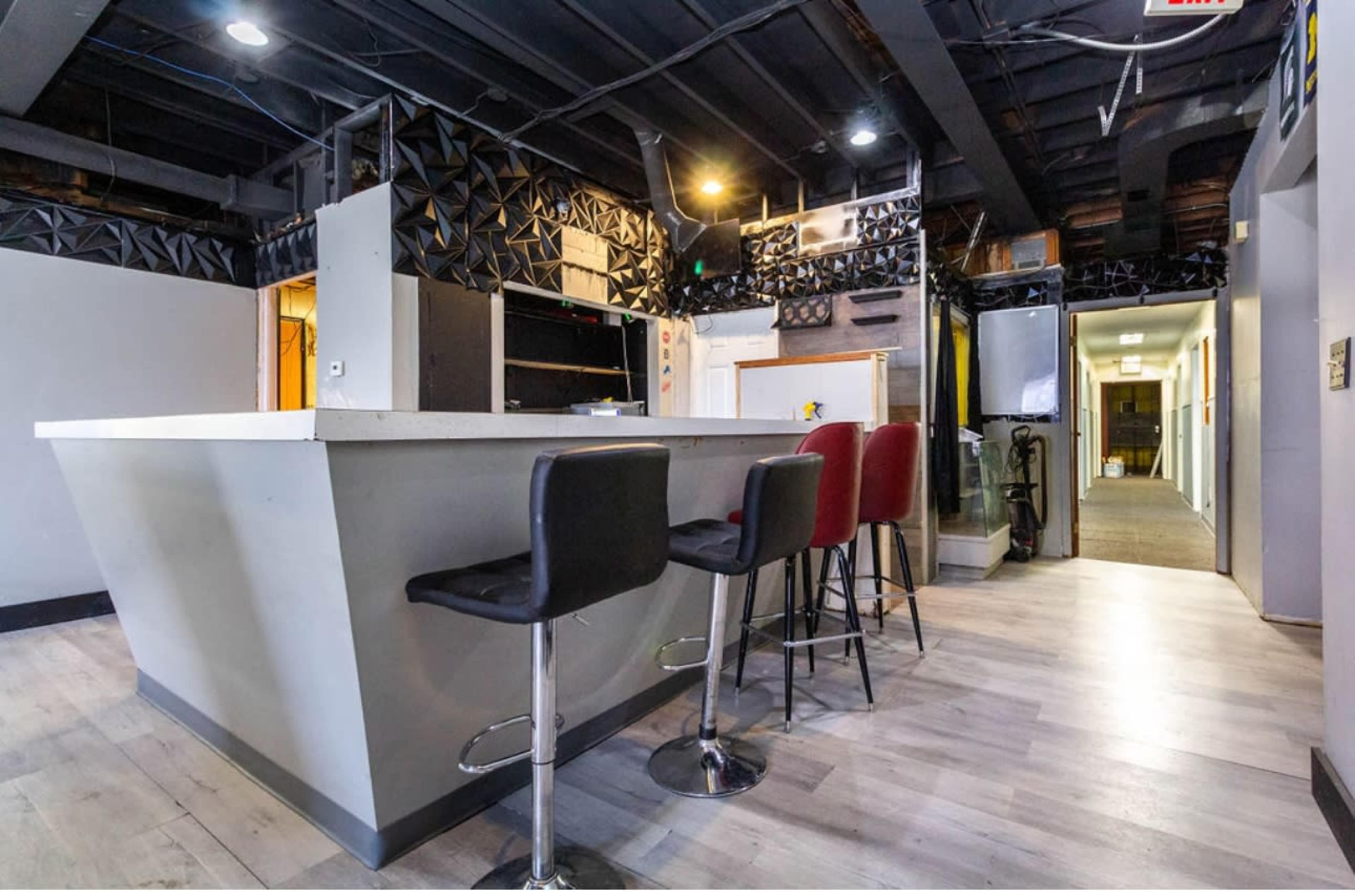 The image shows a modern bar area with a white countertop and several high stools, leading to a corridor with doors on either side.