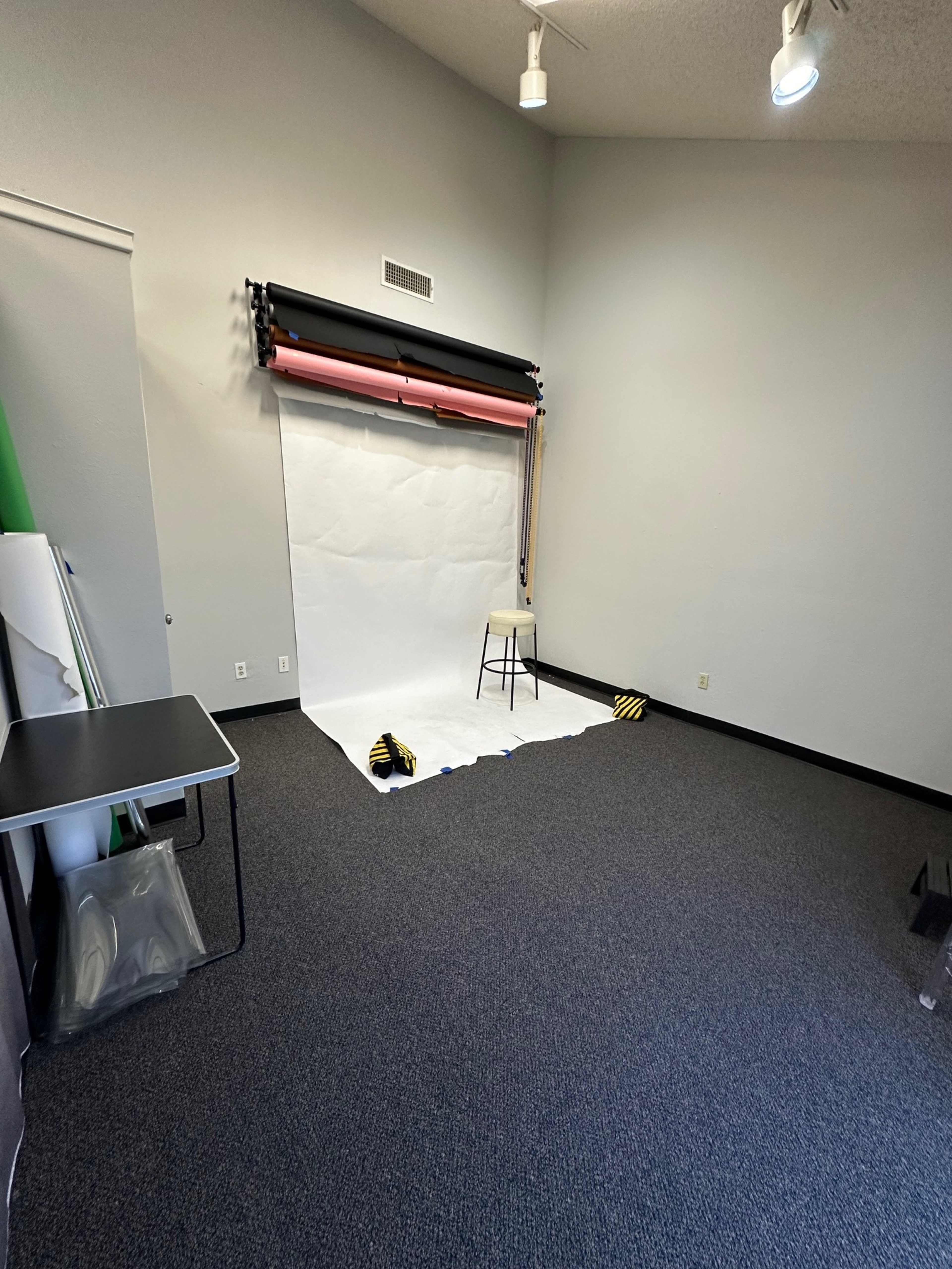 The image shows a photography studio setup with a white backdrop, a stool, and rolled colored backdrops on the wall.
