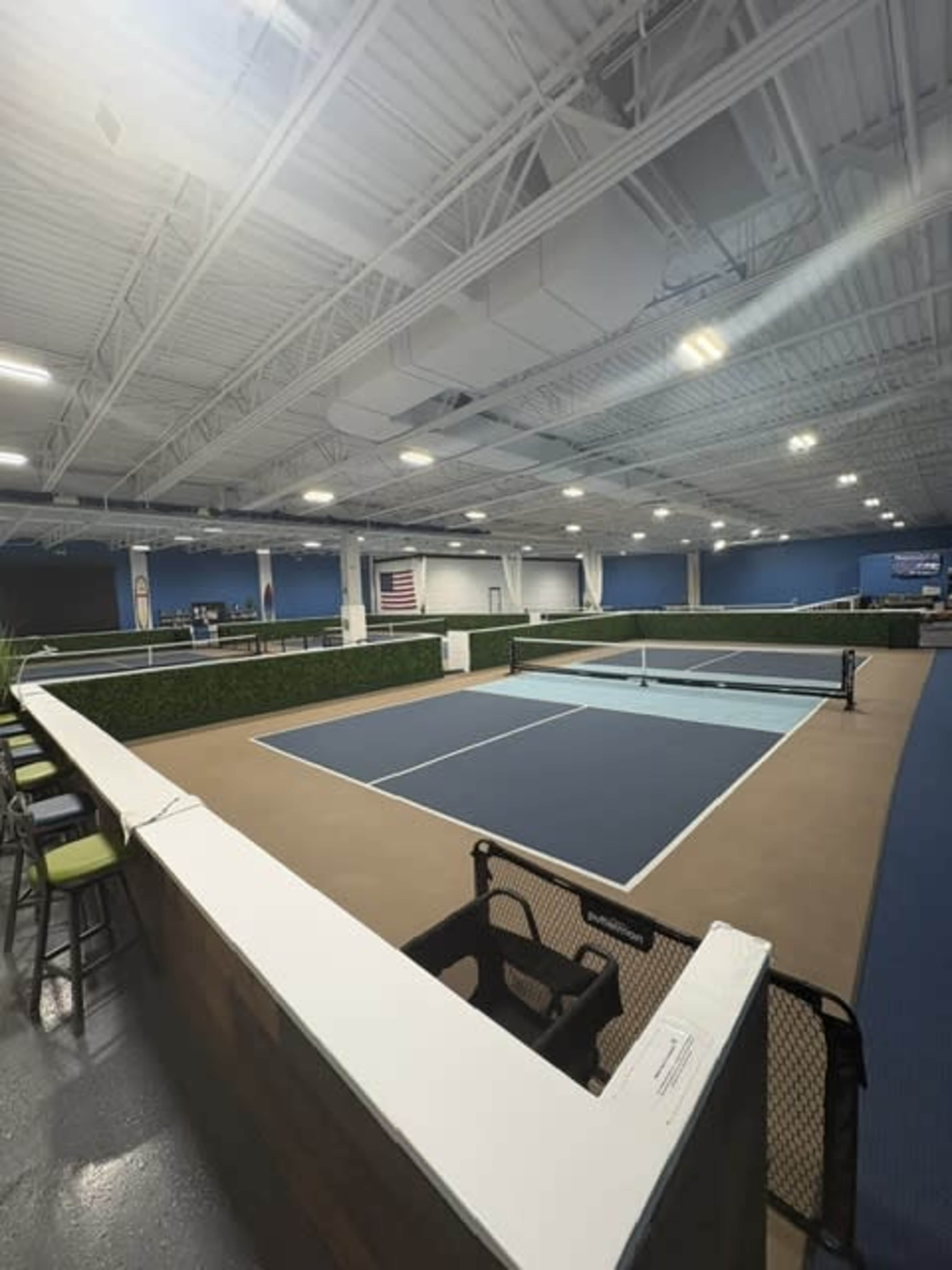 The image shows a spacious indoor tennis facility featuring two blue courts, seating areas, and a flag displayed on the wall.