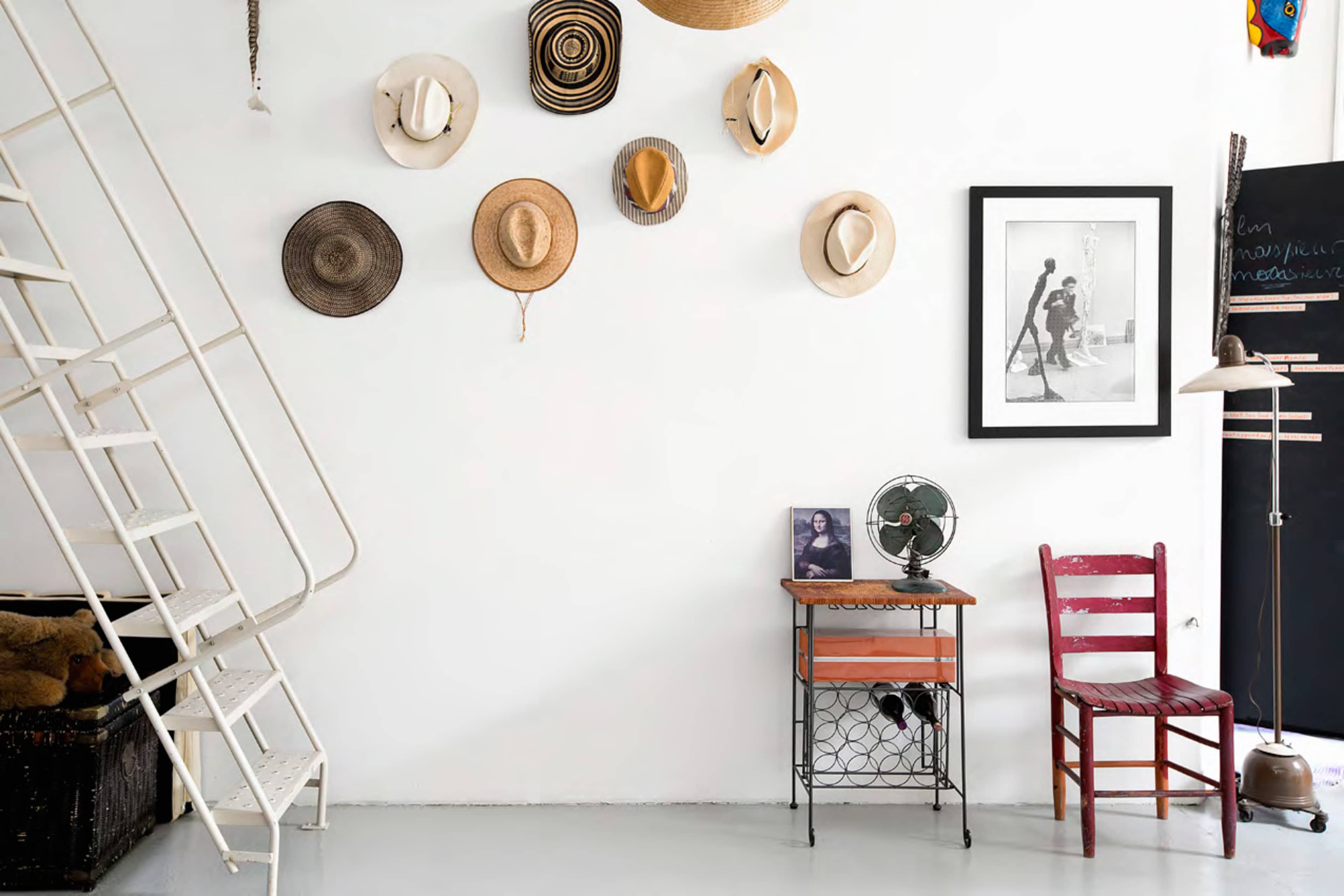 The scene features a white wall adorned with various hats, a framed photo, a red chair, a small table, and a metal fan, alongside a white staircase.