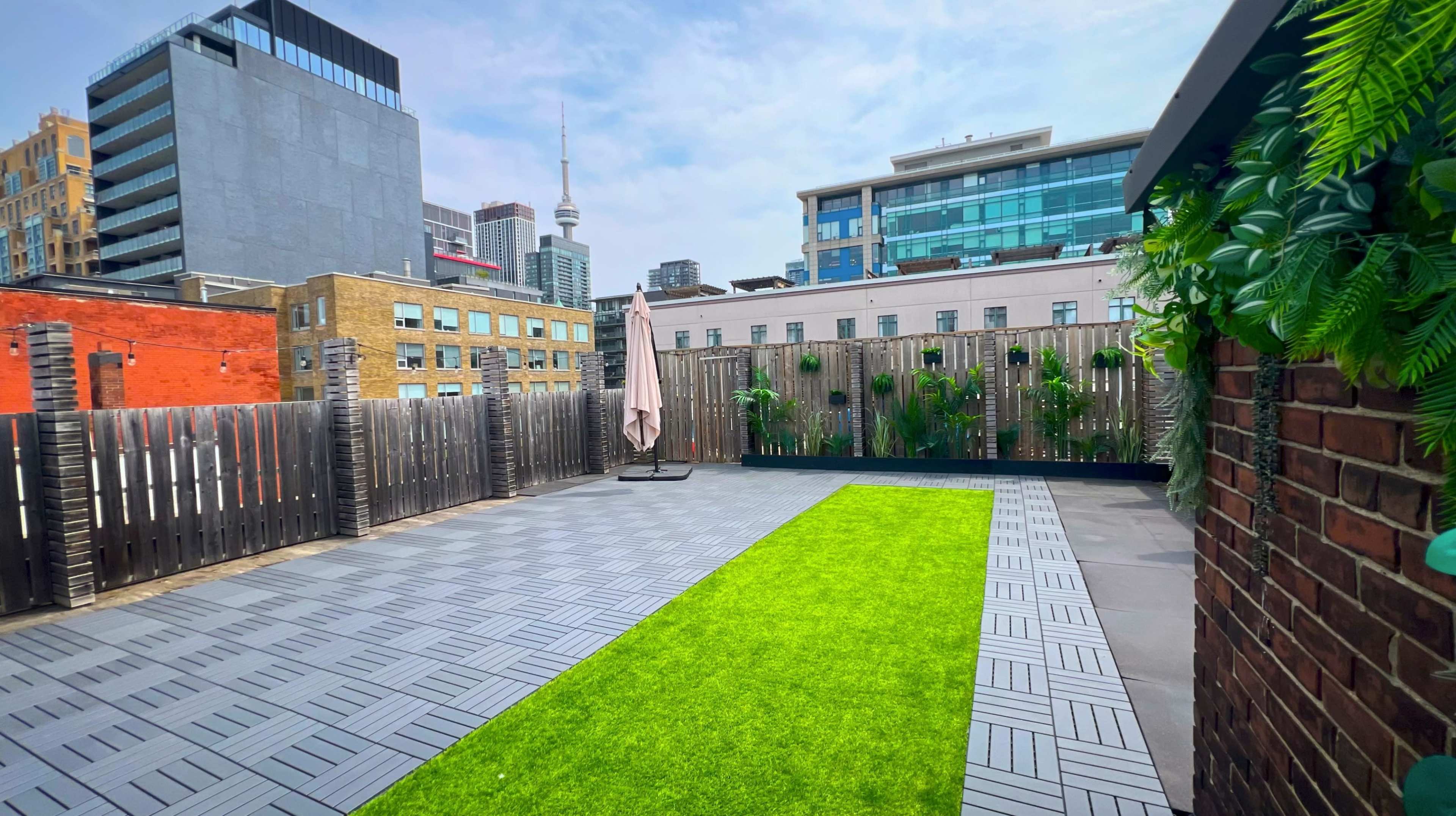 The image shows a rooftop terrace with artificial grass, surrounded by wooden fences and city buildings in the background, including a visible tower.
