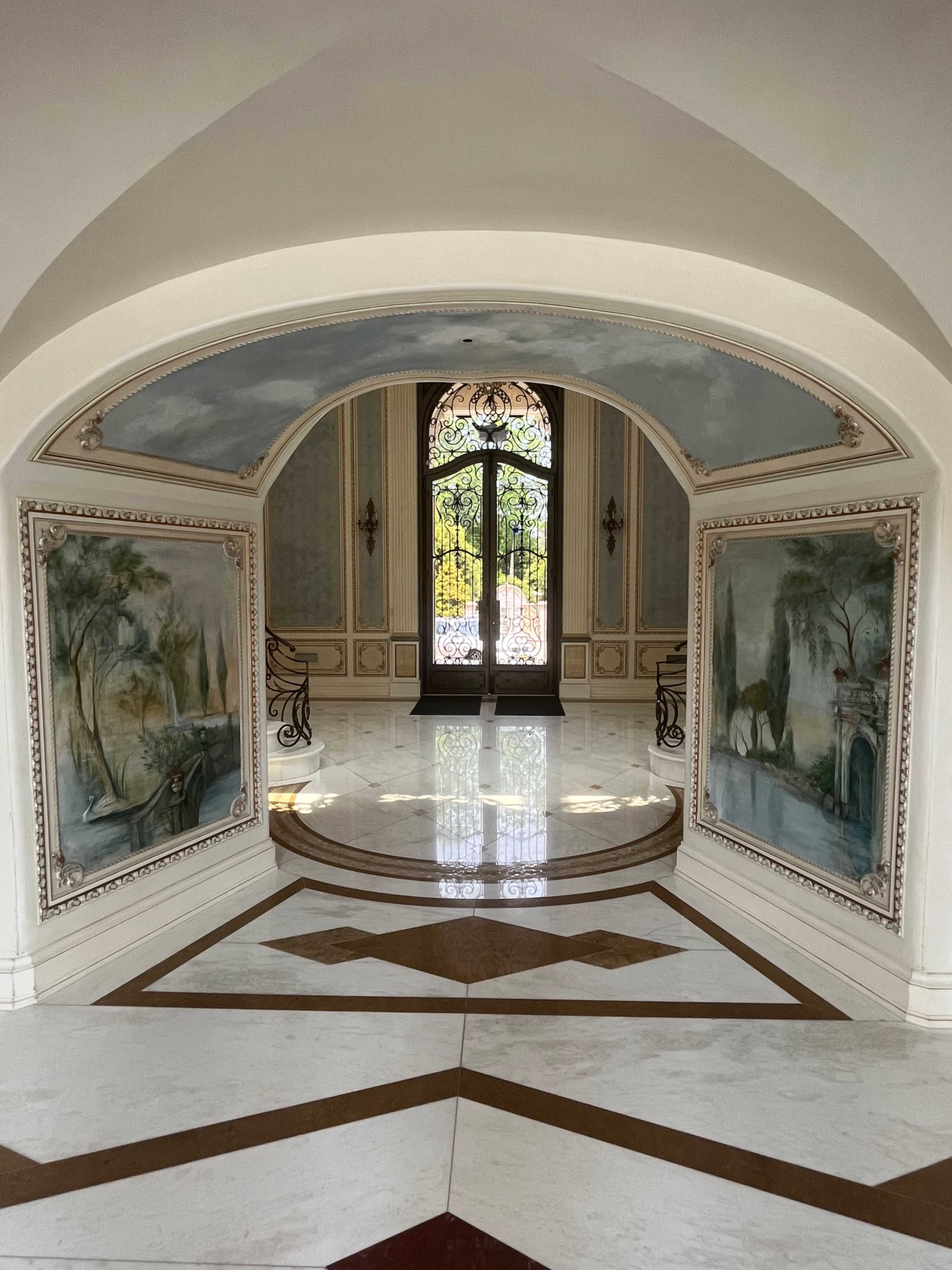 An elegant foyer with arched ceilings, decorative paintings on the walls, and a large, ornate door at the far end leading outside.
