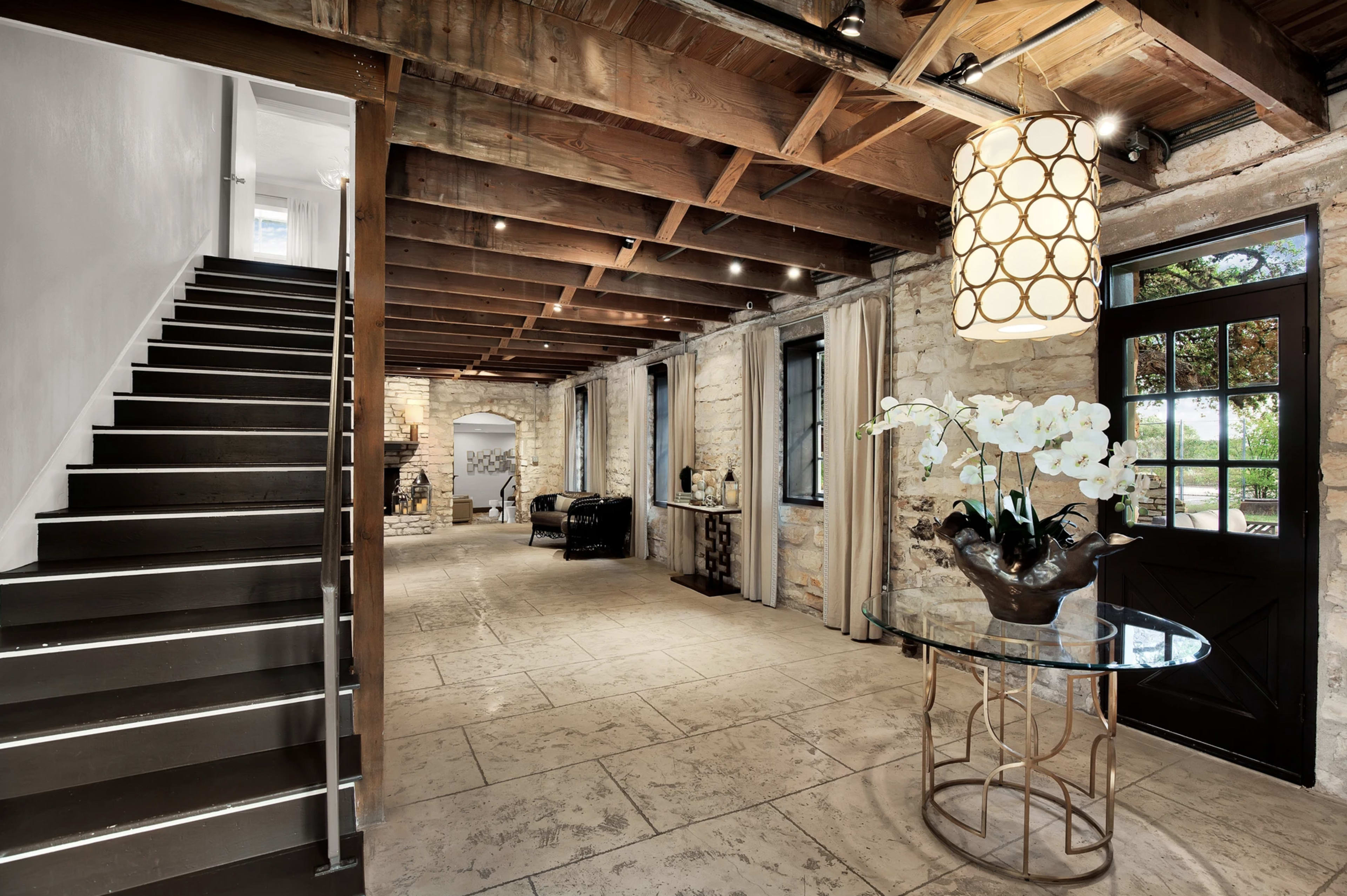 The image shows a modern entryway with a stairway, stone walls, and a glass-topped table featuring a vase of white flowers.
