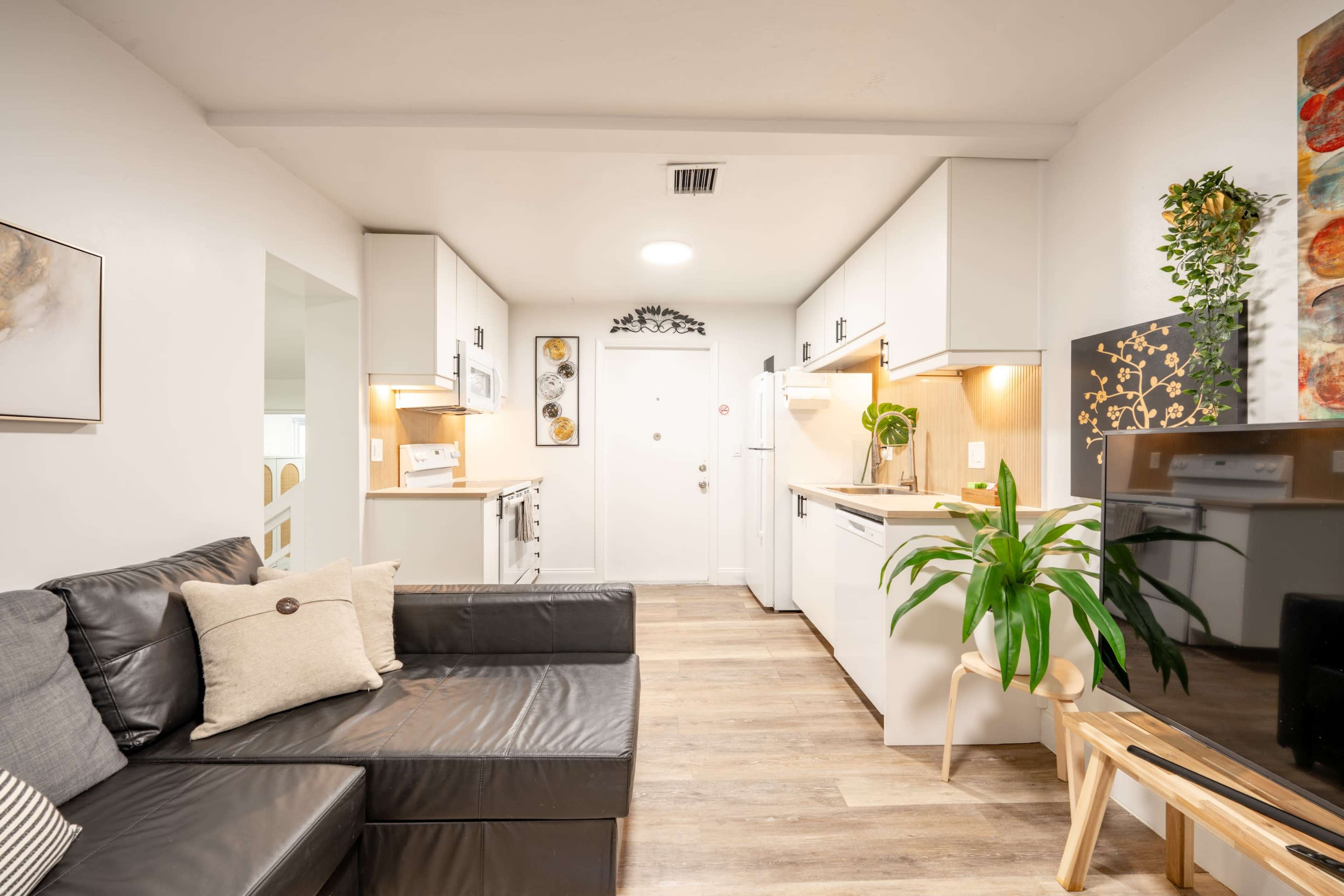 The image shows a modern open-concept living space featuring a black leather sofa, a kitchen with light cabinets, and wooden flooring.
