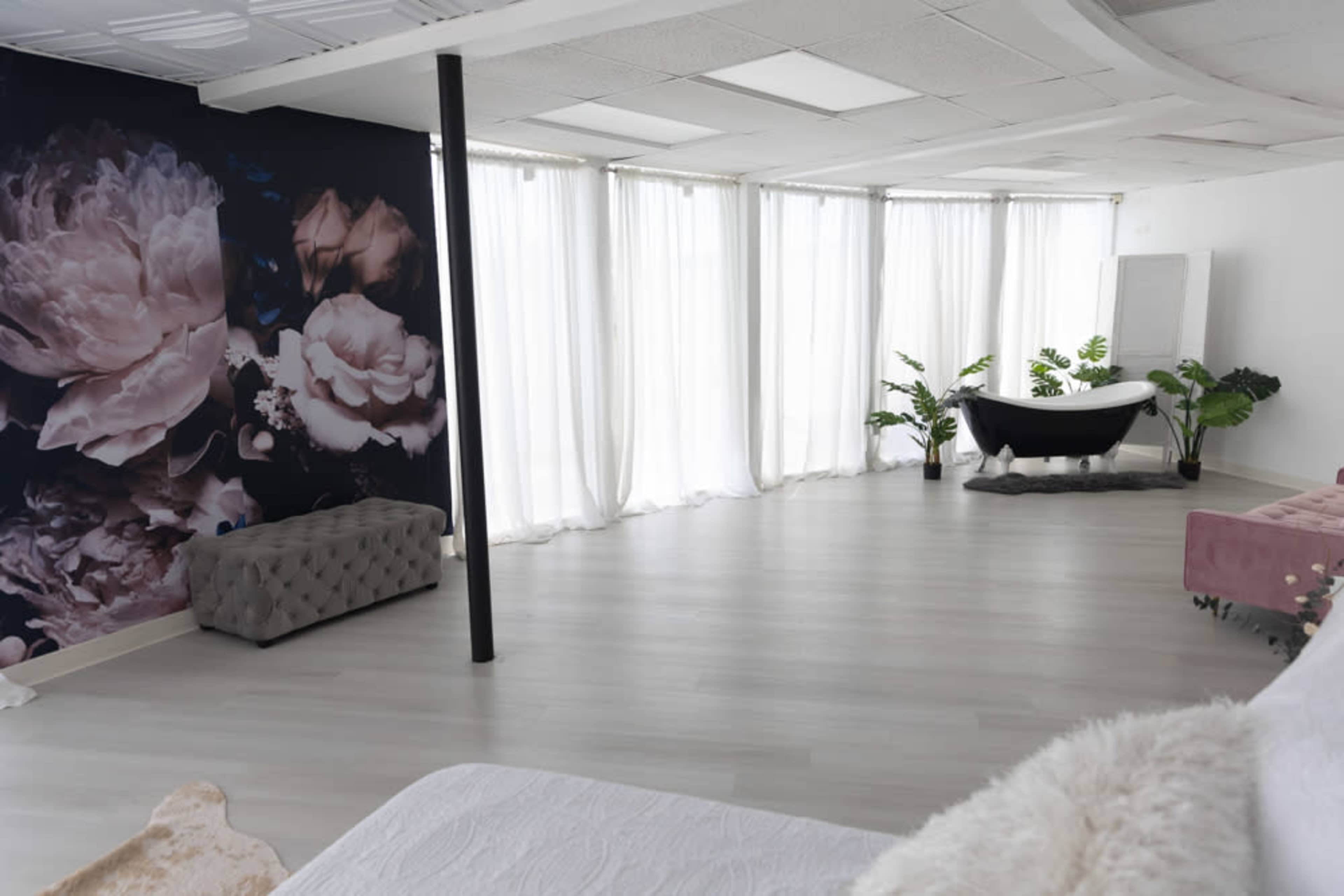 A spacious room with large windows covered by sheer curtains, a bathtub in the corner, a floral wall mural, and minimal furnishings including a tufted bench and a pink sofa.