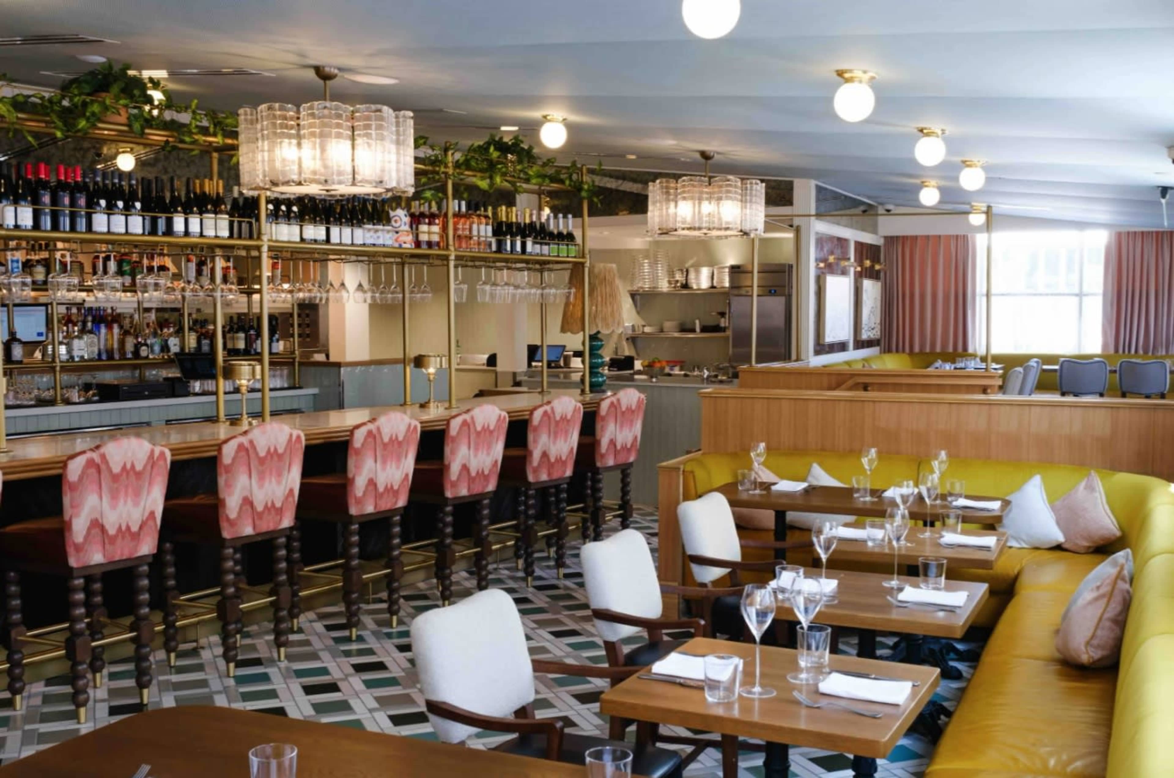 The image shows a modern restaurant interior with a bar, wooden tables, and a mix of seating options, including yellow couches and patterned chairs.