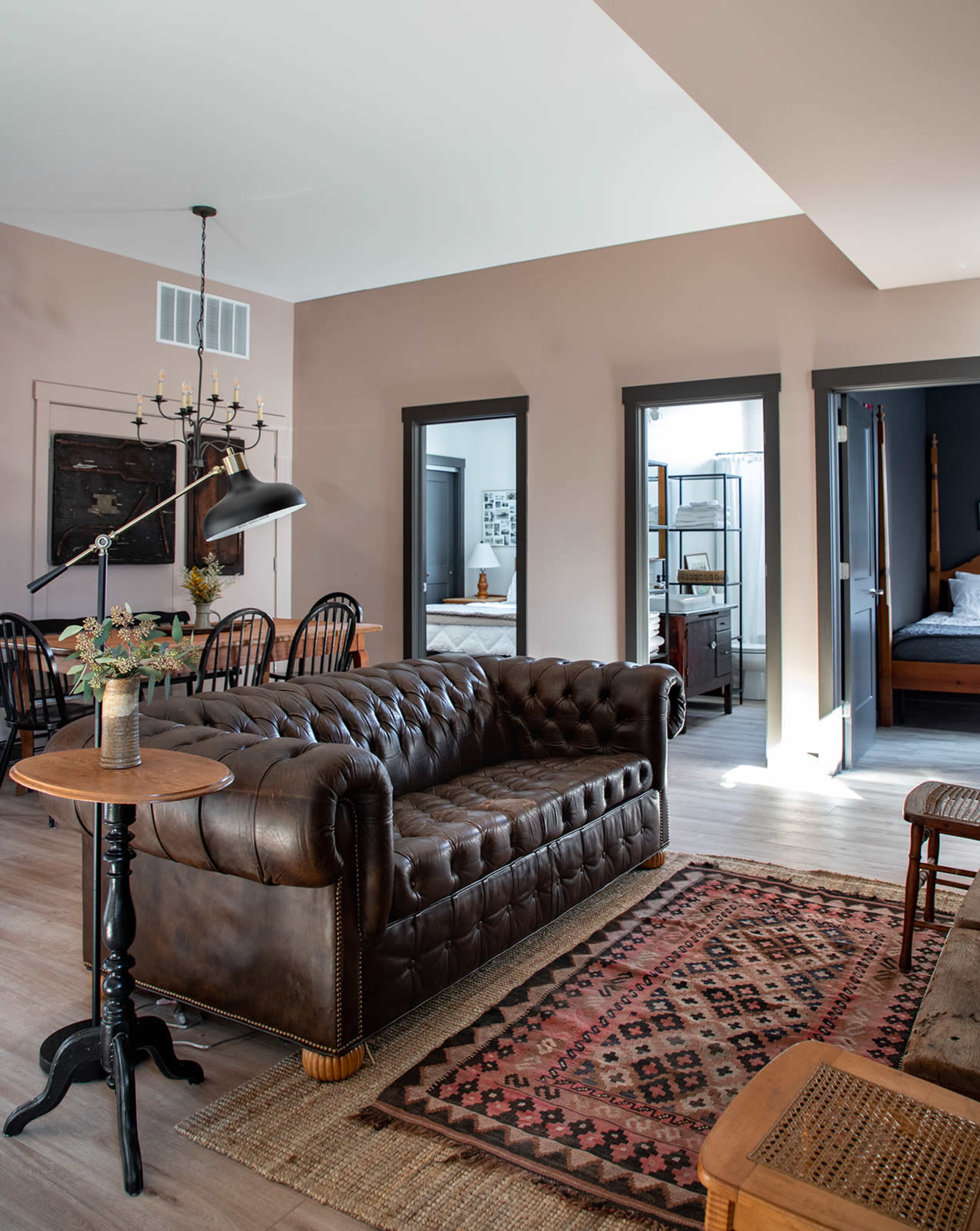 A brown leather couch is positioned in a well-lit living room that connects to a dining area and several doorways leading to other rooms.