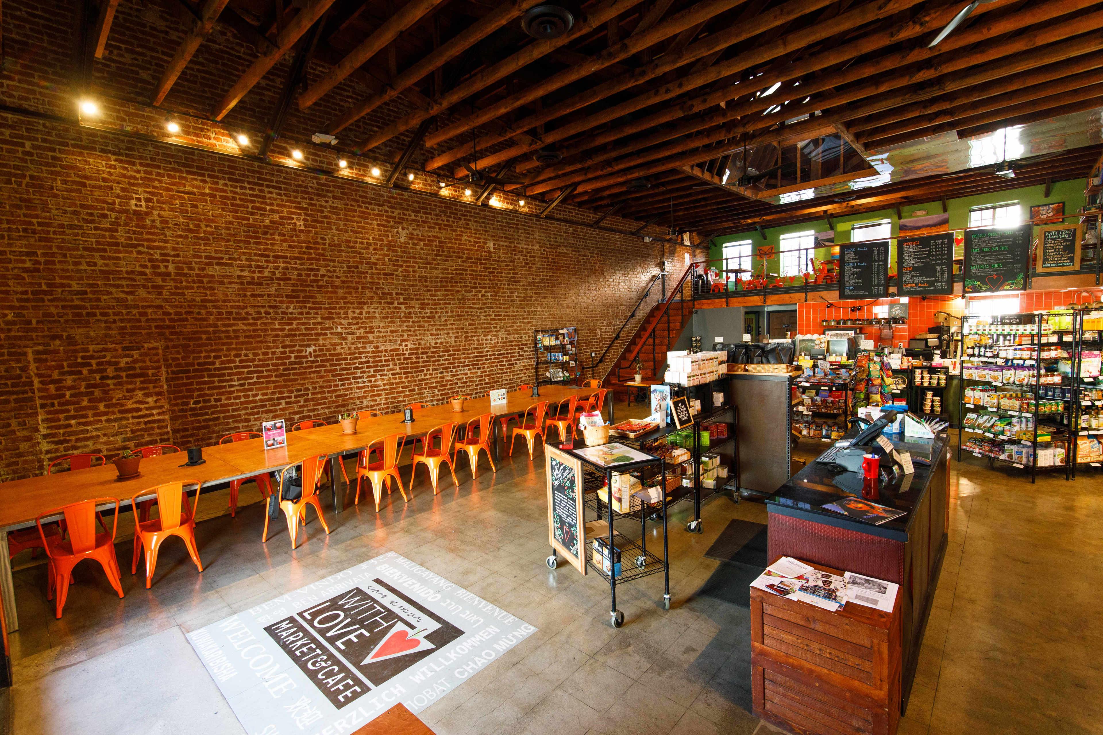 A spacious café features wooden tables and orange chairs along a brick wall, with shelves of products and a service counter in the foreground.