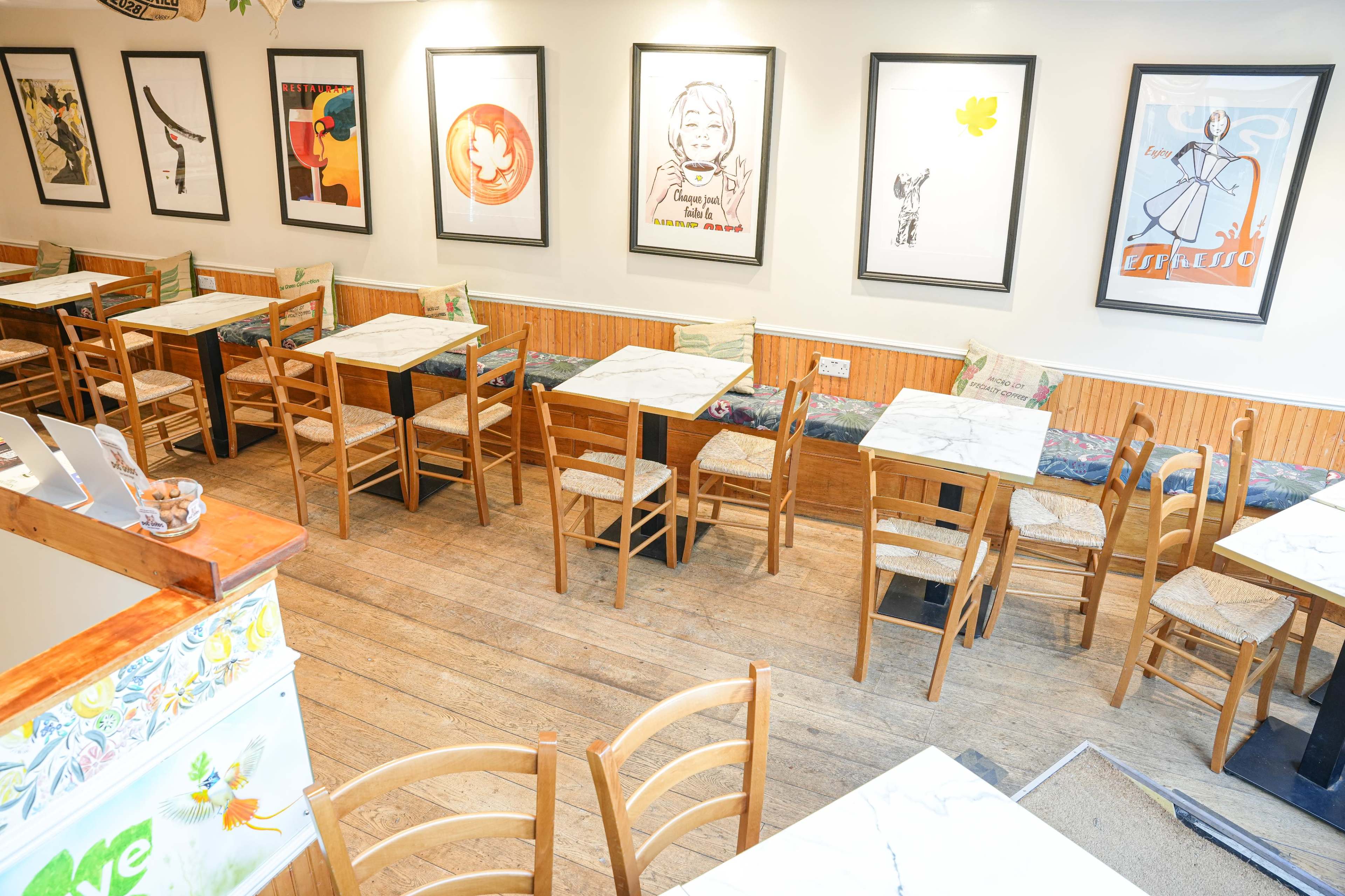 A warmly lit cafe with wooden tables and chairs arranged neatly, framed artworks on the walls, and a visible counter area.