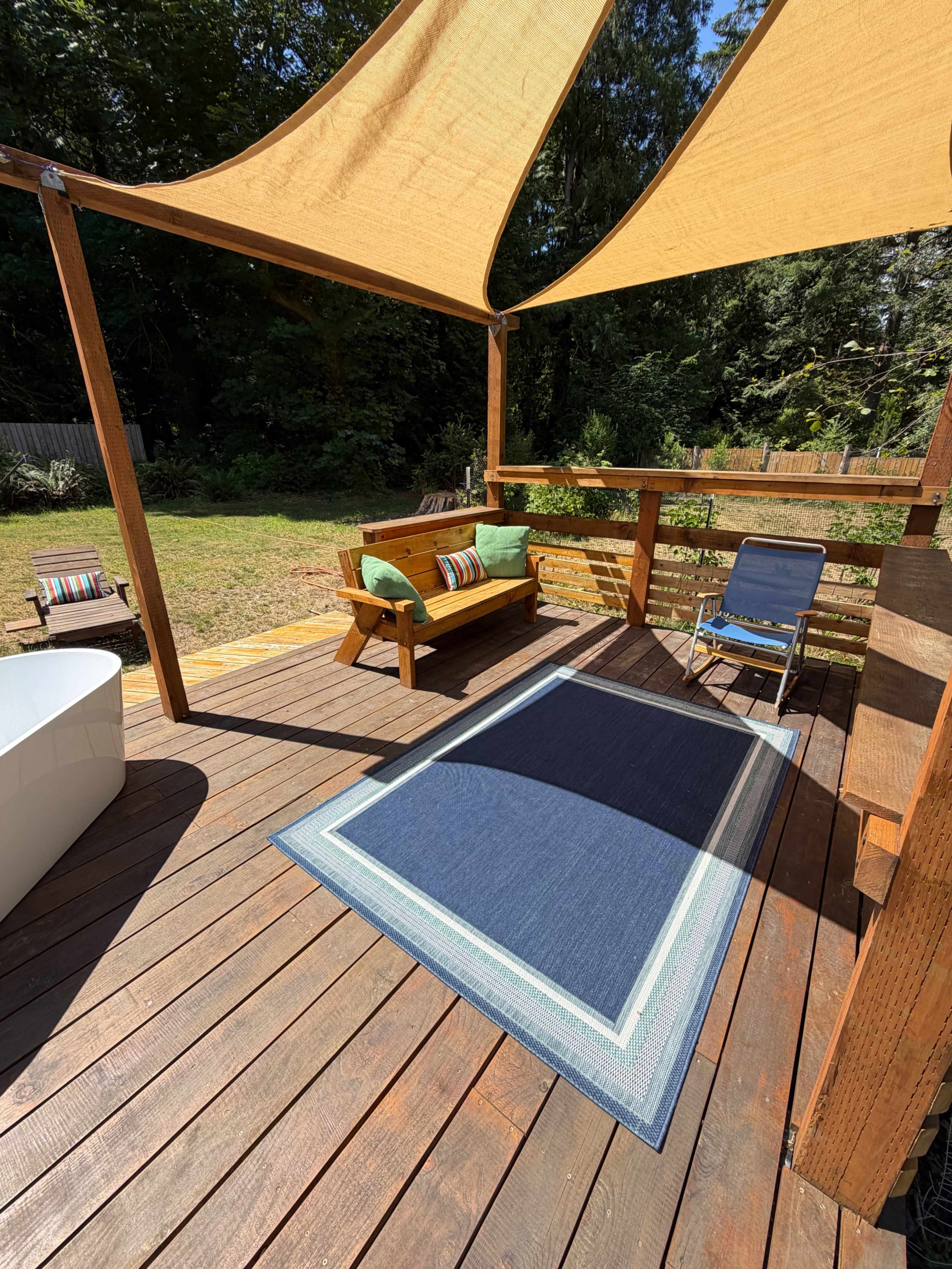 The image shows a deck area with a large shade sail, a wooden bench with cushions, a folding chair, and a blue rug, surrounded by trees and grass.
