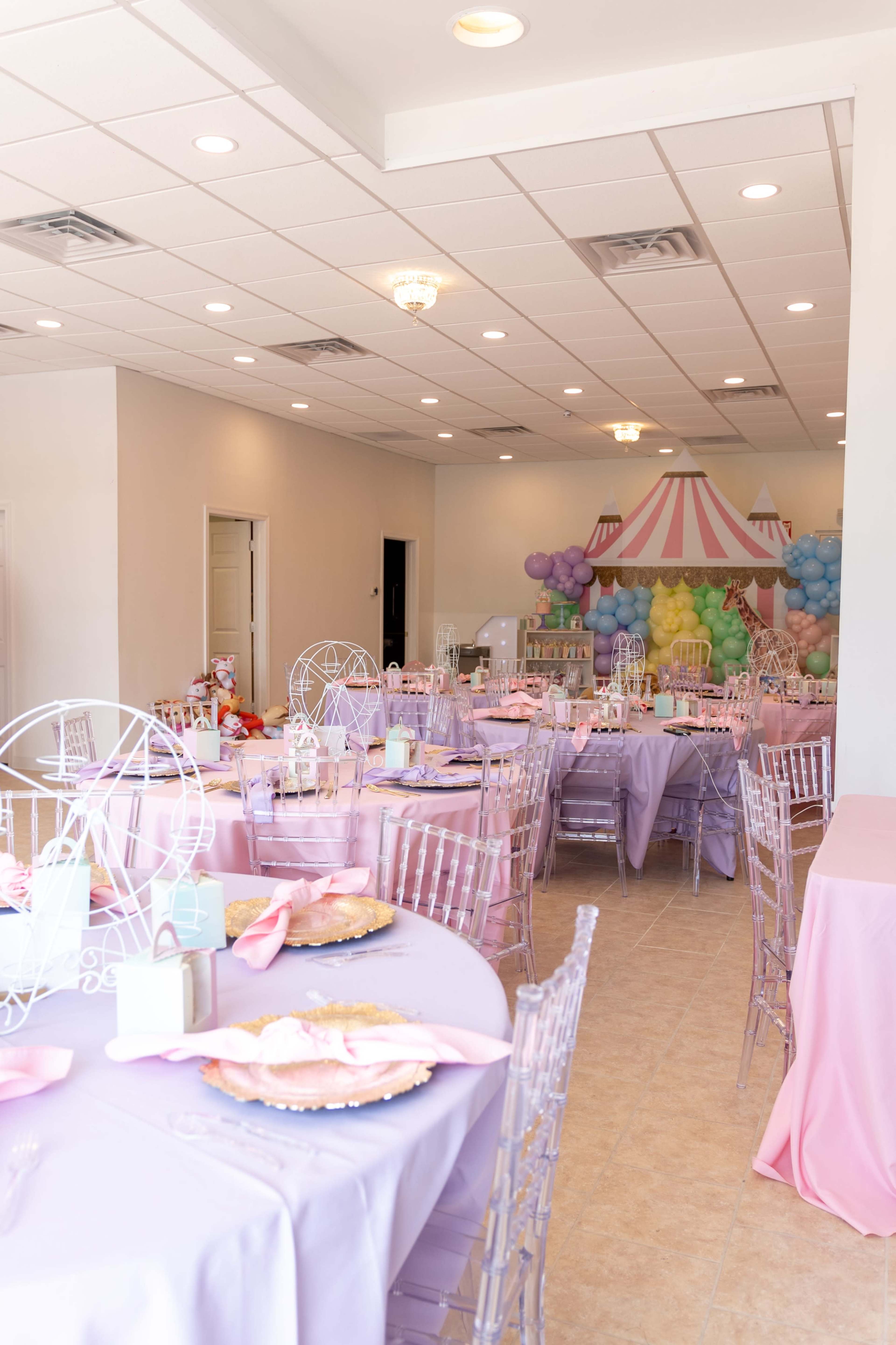 The image shows a banquet hall set up for an event, featuring round tables covered in pastel lavender tablecloths and decorated with pink napkins, alongside a backdrop of colorful balloons and a striped tent motif.