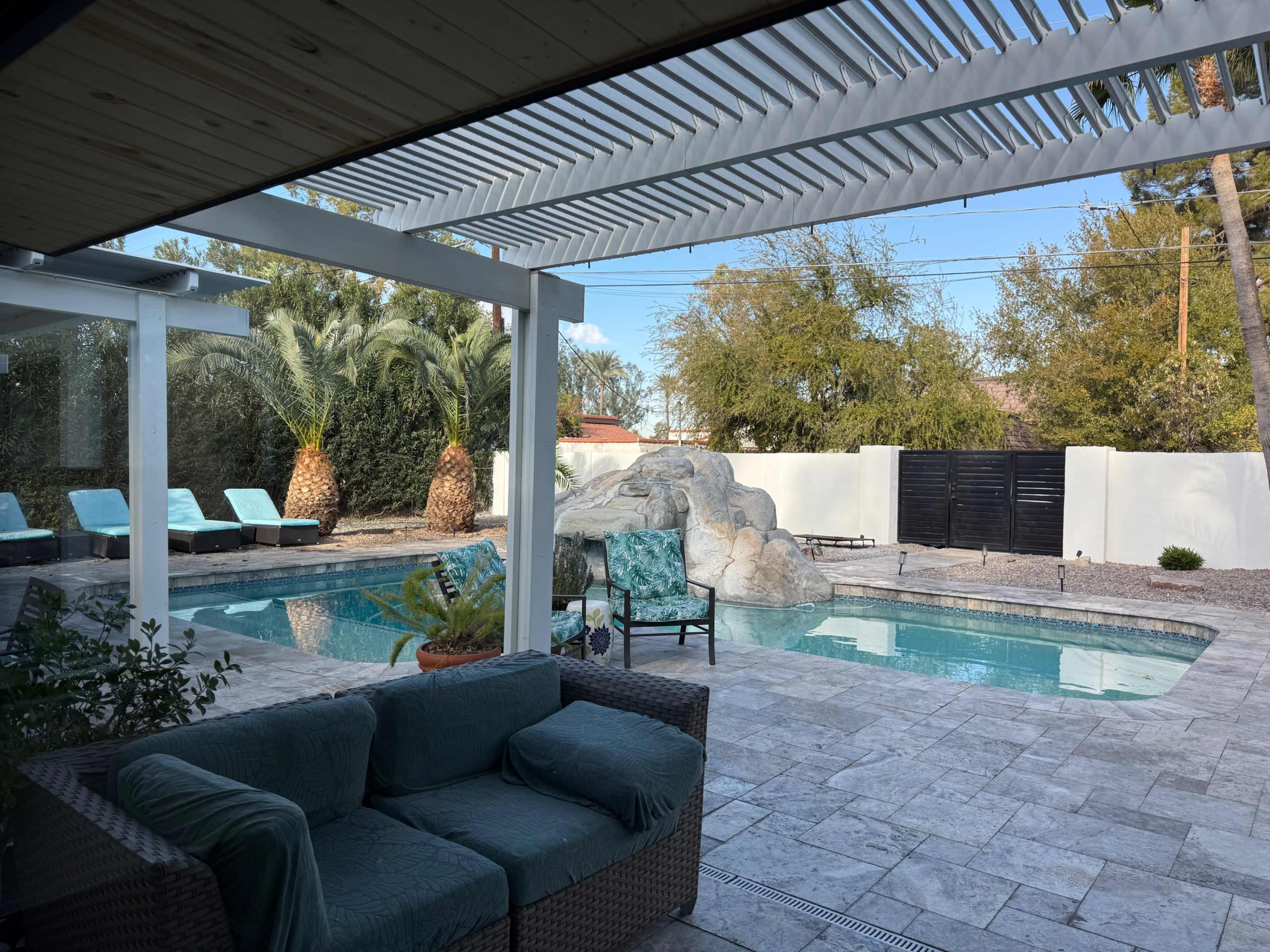 The image shows a backyard with a swimming pool surrounded by lounge chairs, palm trees, and a rock feature, viewed from a shaded patio area.