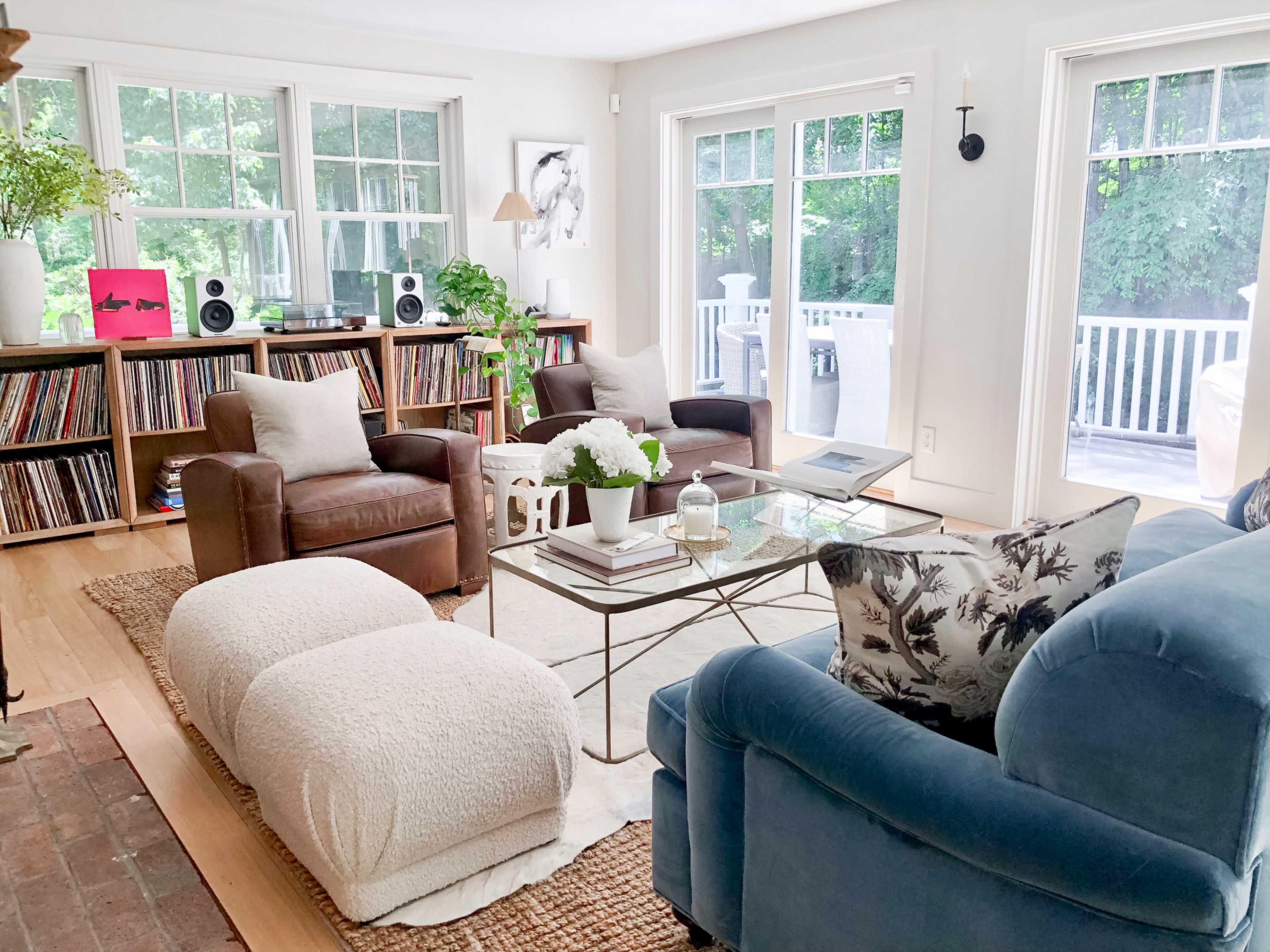 A bright living room with a blue sofa, two brown armchairs, a glass coffee table, and large windows overlooking a deck.