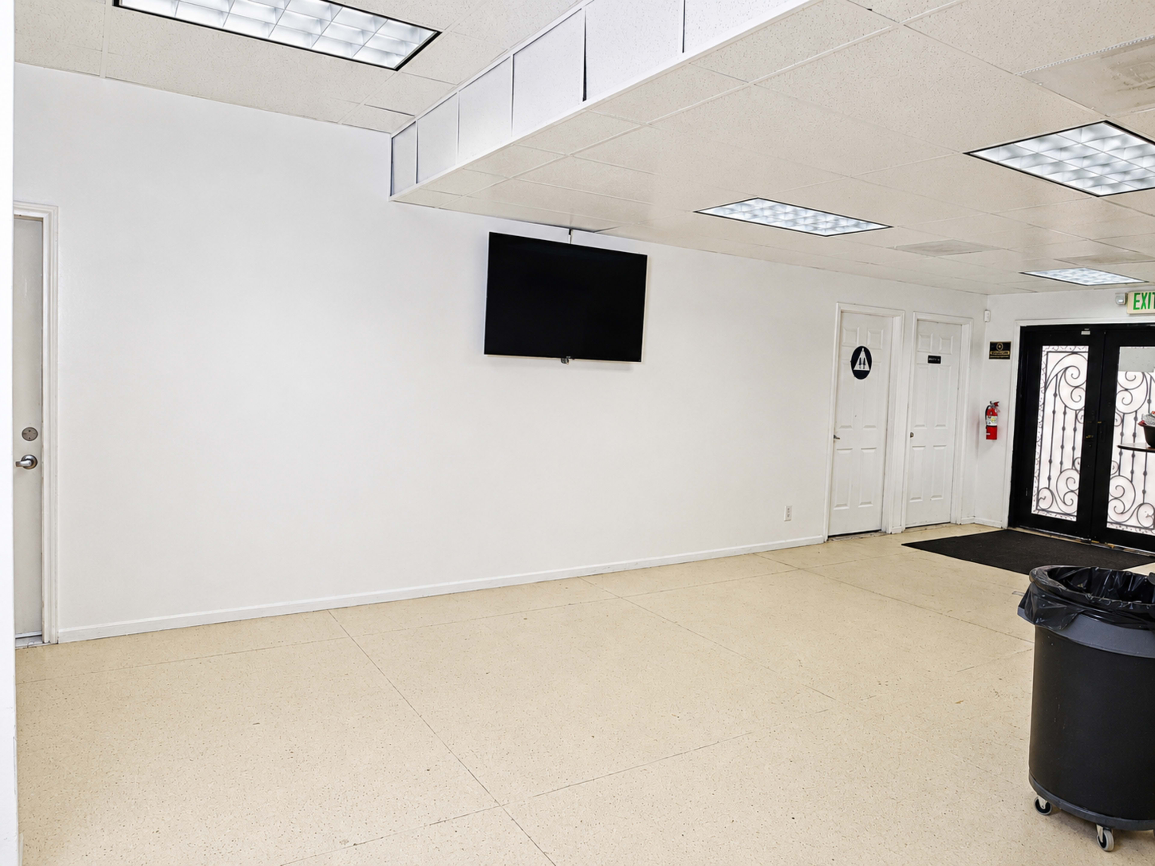 The image shows a spacious, empty room with a white wall, a mounted television, and a door leading outside, alongside a trash bin in one corner.