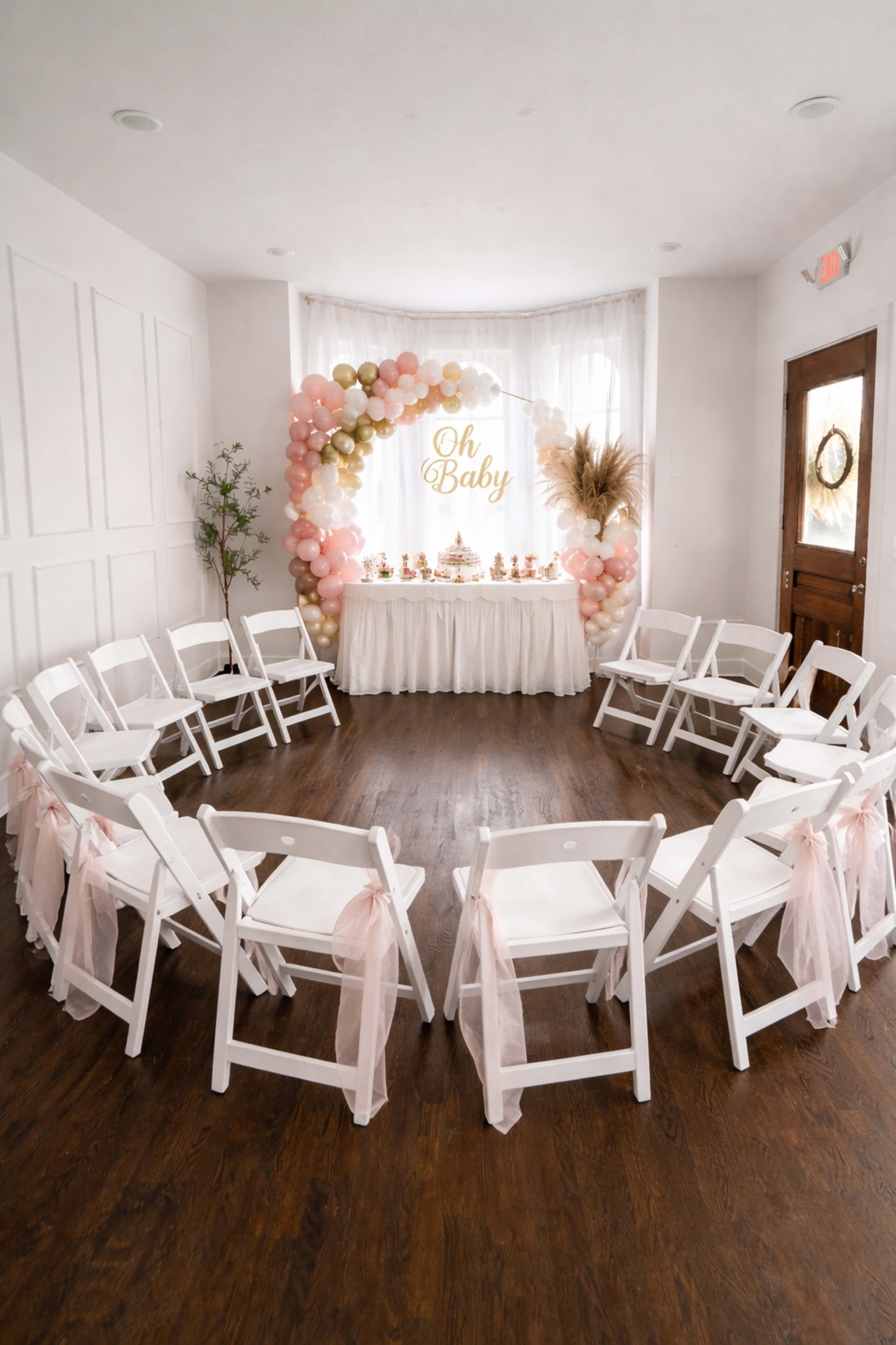 A cozy indoor space is arranged with white chairs in a circular formation around a decorated table featuring a balloon arch and a sign reading "Oh Baby."