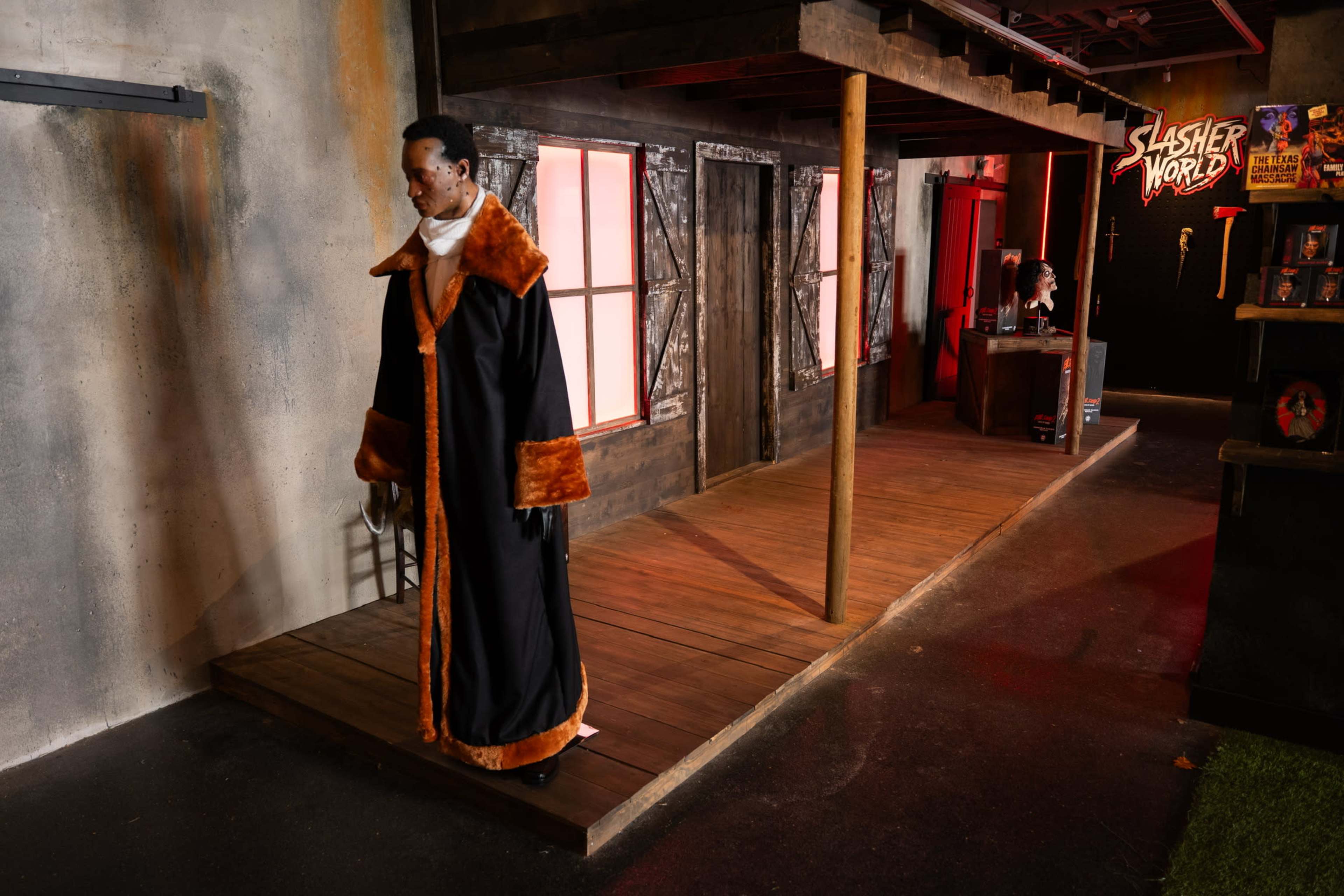 A mannequin dressed in a long black coat with orange trim stands on a wooden platform in a dimly lit room decorated with horror-themed elements.