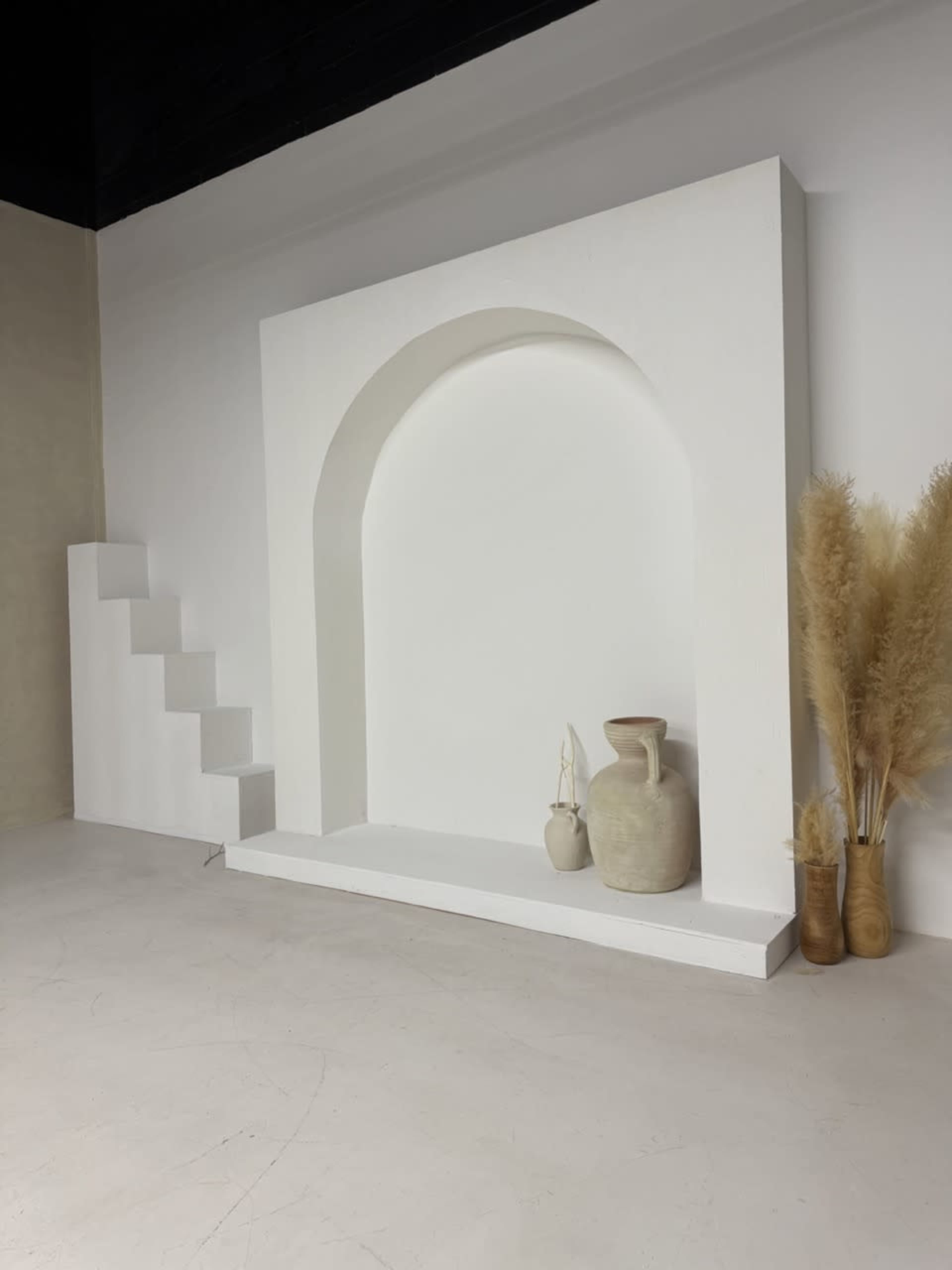A minimalist interior scene features a white, arch-shaped wall niche flanked by two decorative vases and dried plants, with a set of white stairs on the left.
