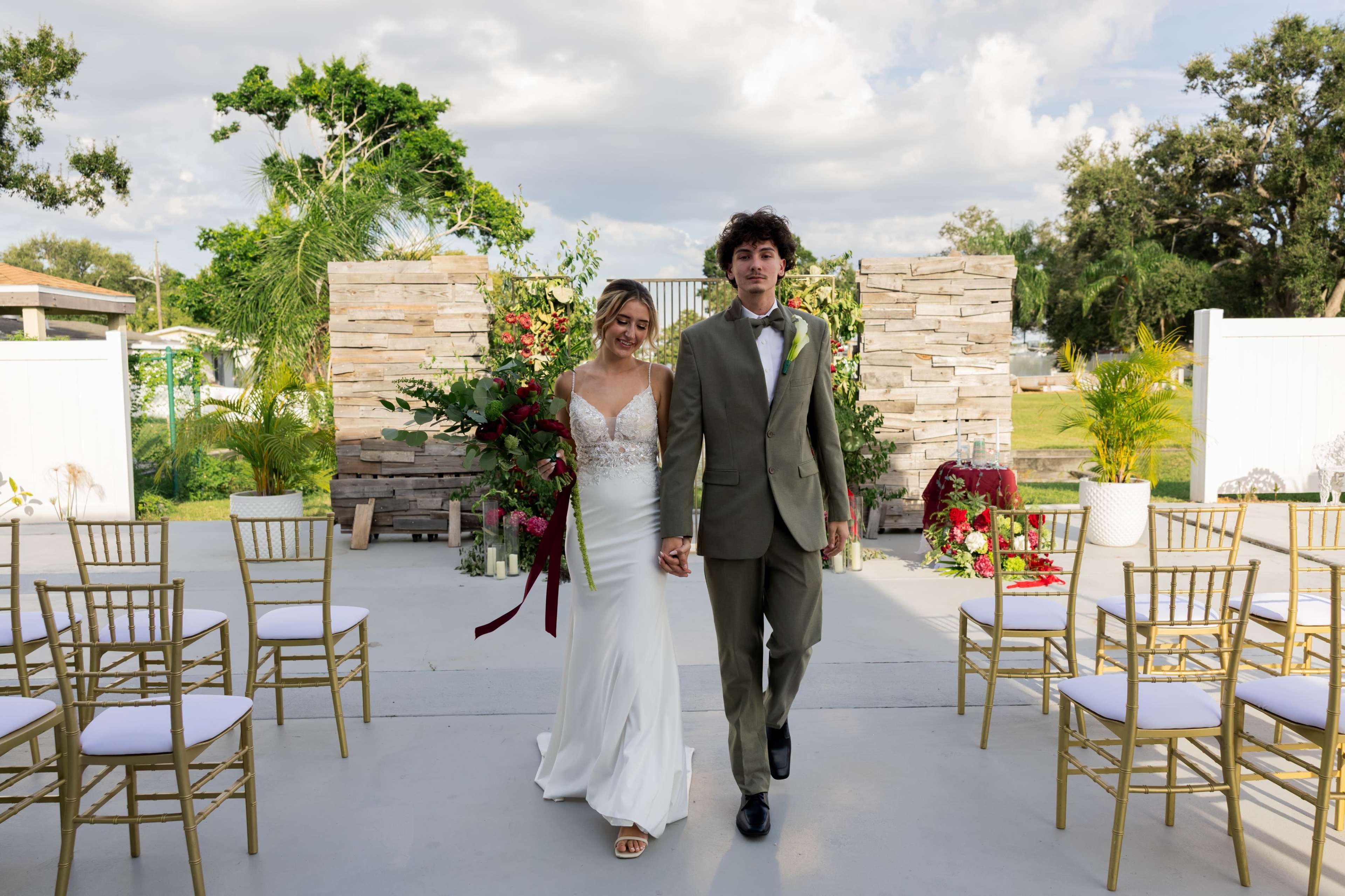 A bride in a white gown and a groom in a green suit walk hand in hand down a decorated outdoor aisle with chairs on either side.