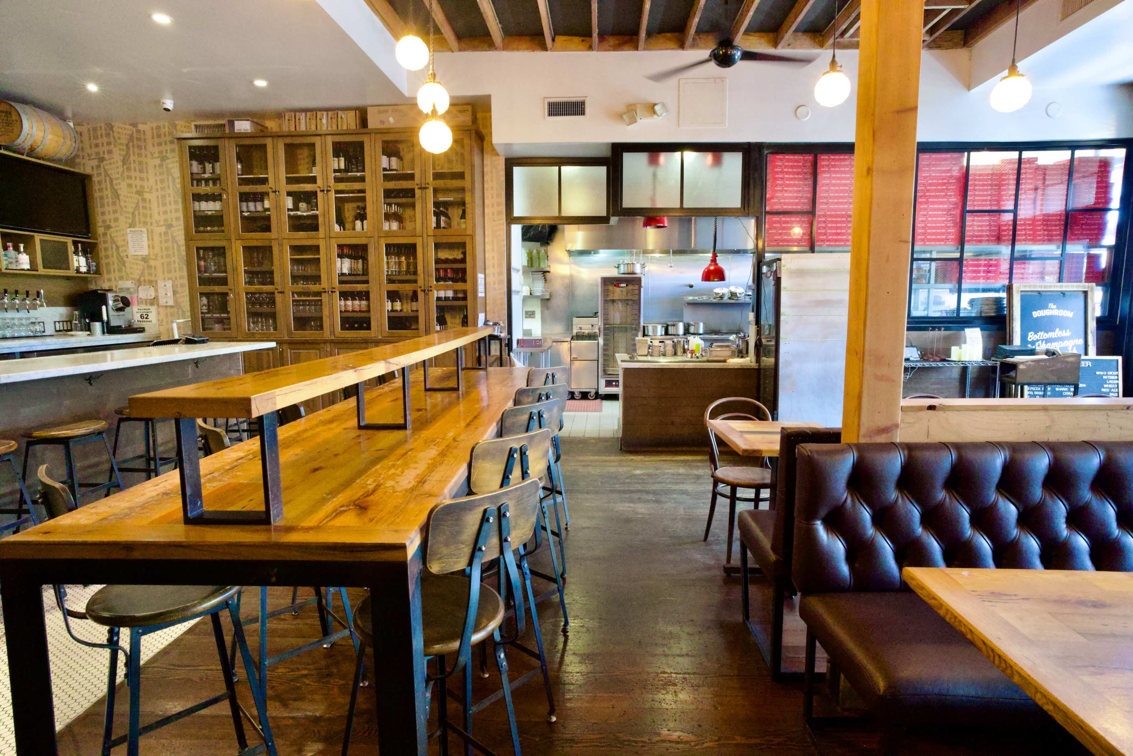 The image shows a modern restaurant interior with wooden tables, metal chairs, and an open kitchen area at the back.