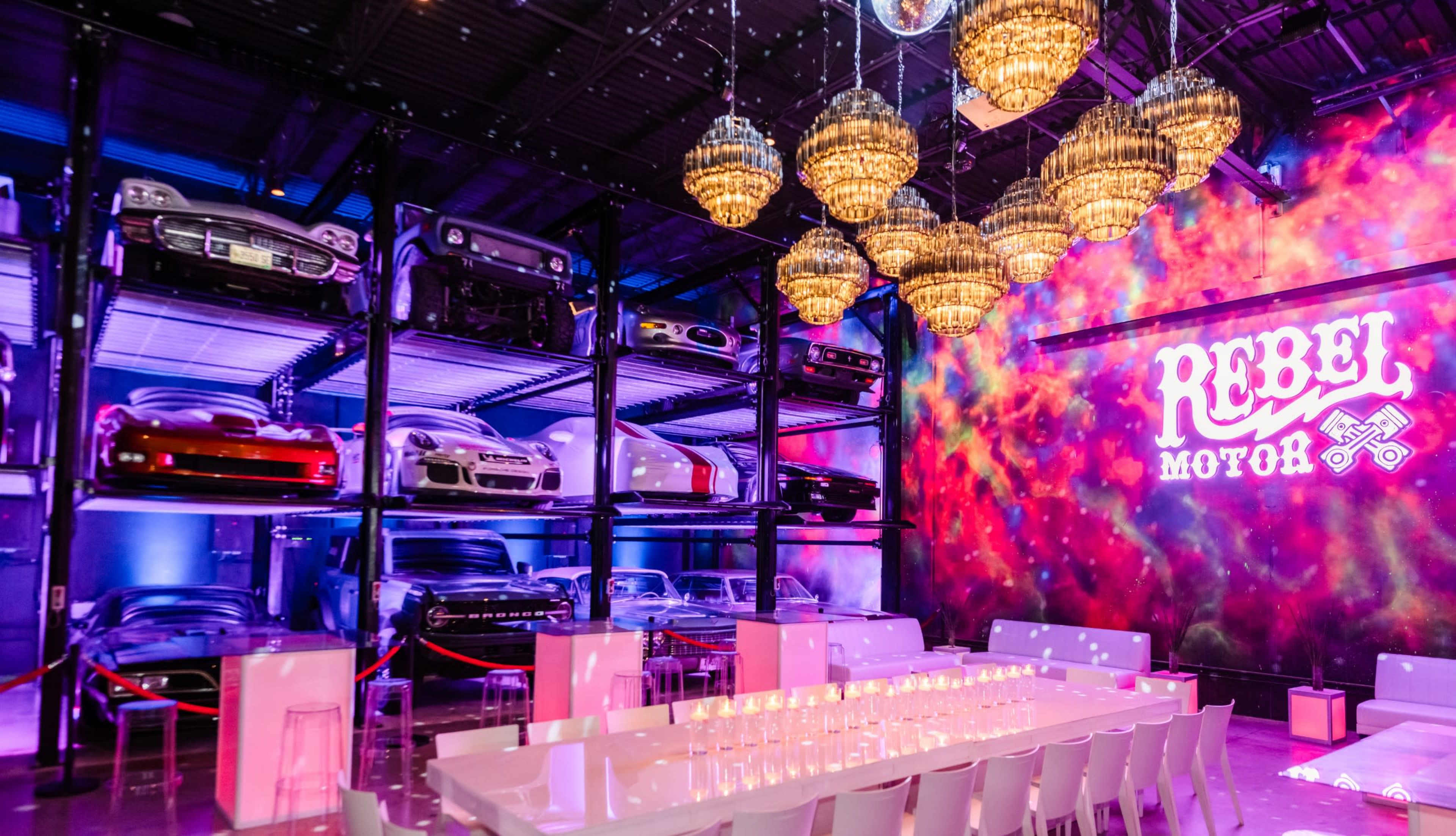A modern event space featuring a row of luxury cars displayed on shelves, with a long table set for dining beneath decorative chandeliers and a colorful wall mural.
