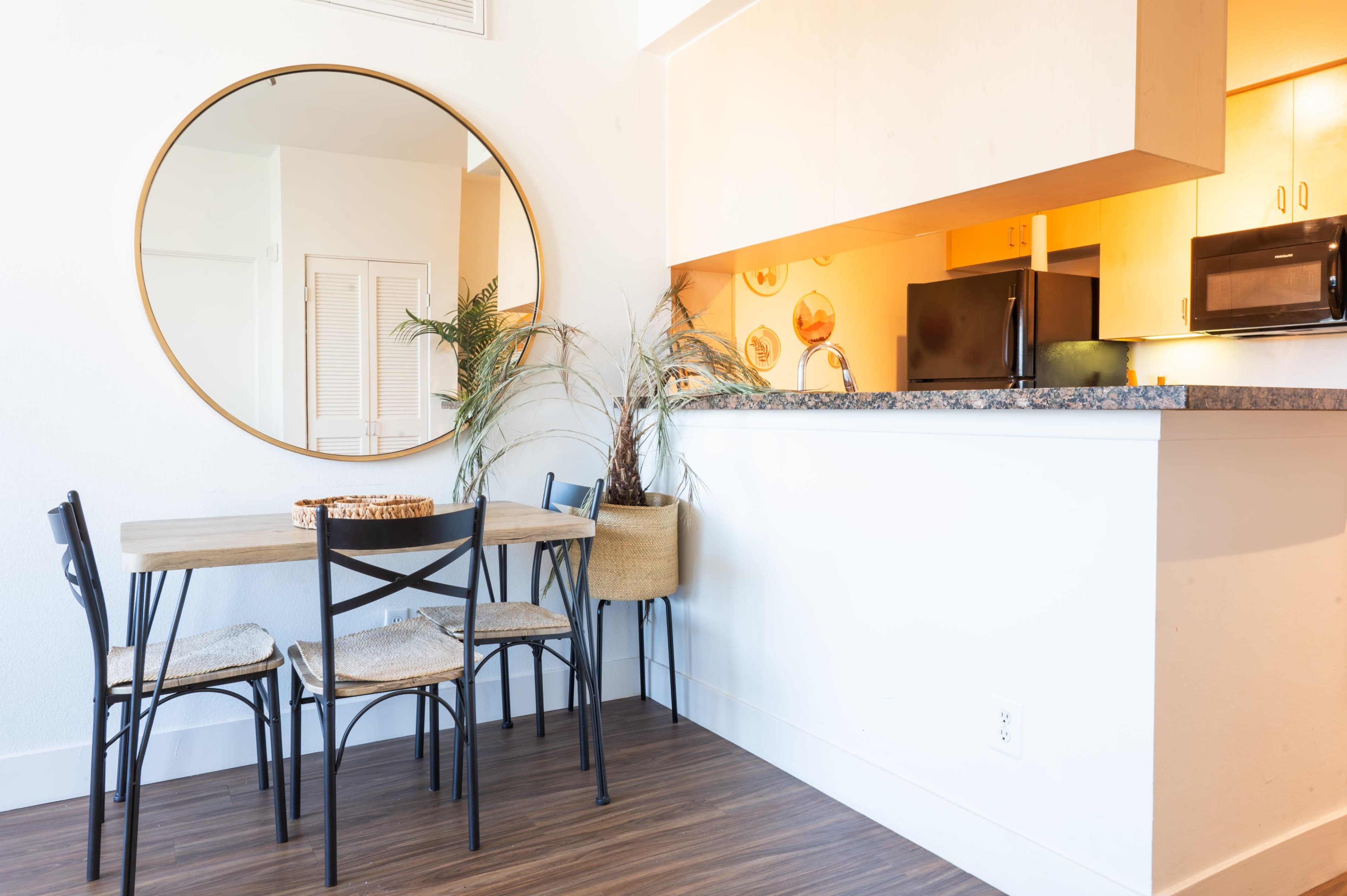 A dining area features a round mirror, a wooden table with four black chairs, and a small kitchen counter with appliances and decorative elements.