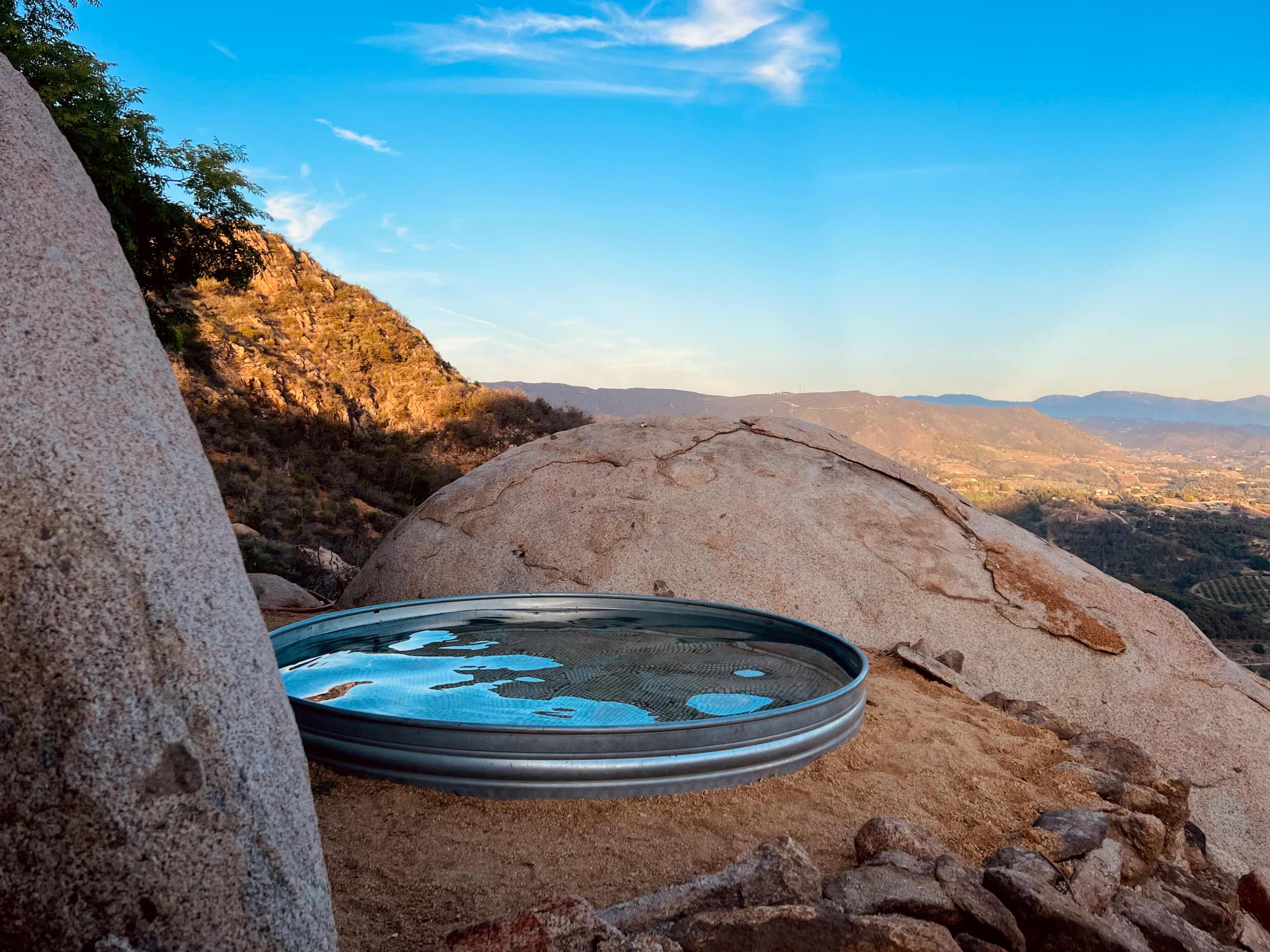 A circular metal dish sits on a rocky surface, reflecting a clear sky and surrounding landscape.