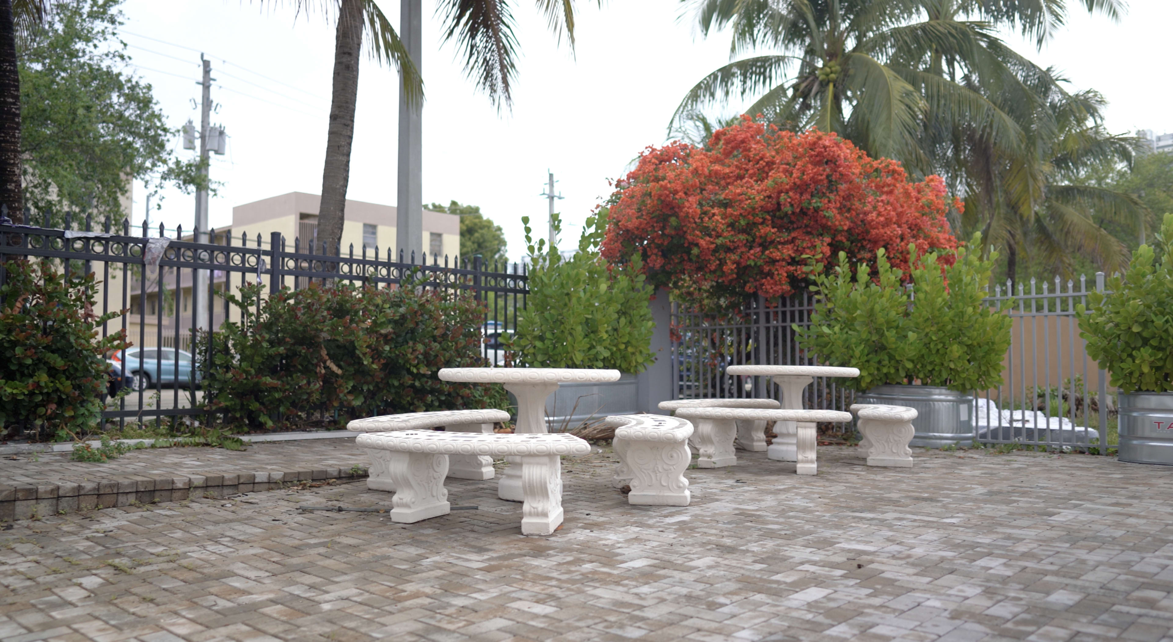 A small outdoor seating area with white stone tables and benches surrounded by greenery and flowering shrubs.