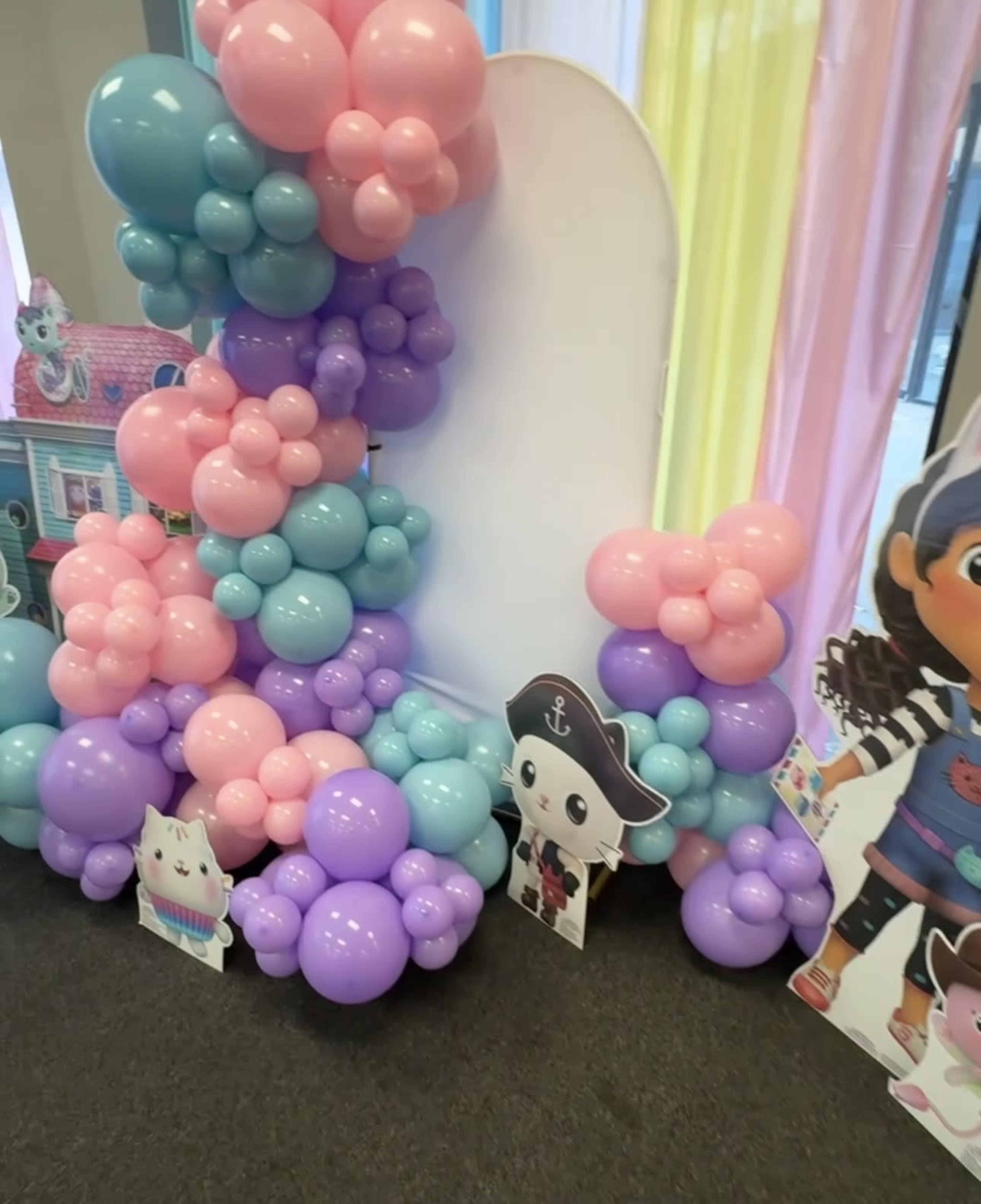 The image shows a colorful arrangement of balloons in pink, blue, and purple, alongside cutouts of cartoon characters, set against a backdrop of pastel drapes.